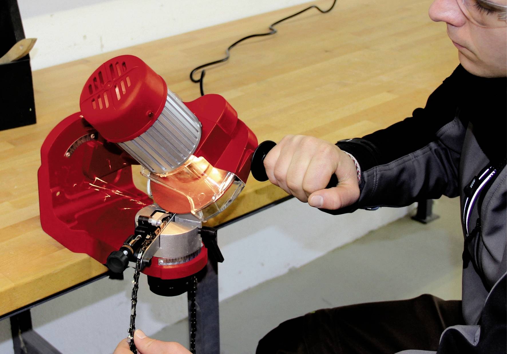 Person benutzt eine rote Schleifmaschine auf einer Werkbank, um eine Kette zu schärfen. Die Person hält die Kette mit einer Hand.