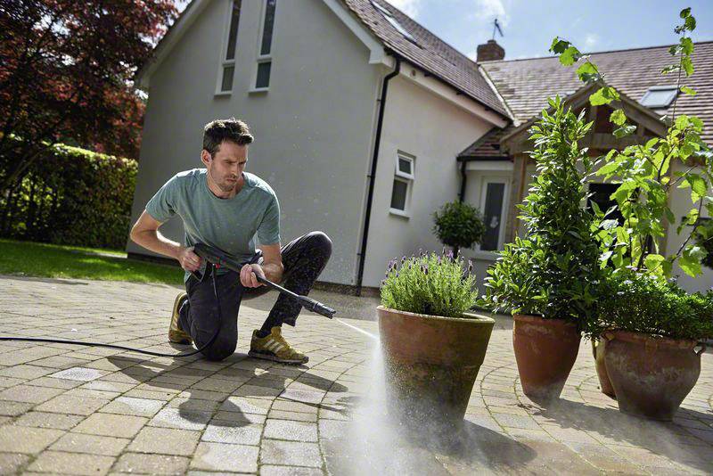 Eine Person reinigt die Terrasse mit einem Hochdruckreiniger neben Pflanzen in Töpfen. Im Hintergrund ist ein Haus zu sehen.