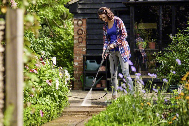 Frau reinigt Terrasse mit Hochdruckreiniger im Garten. Im Hintergrund ein Haus mit Blumen und Pflanzen.
