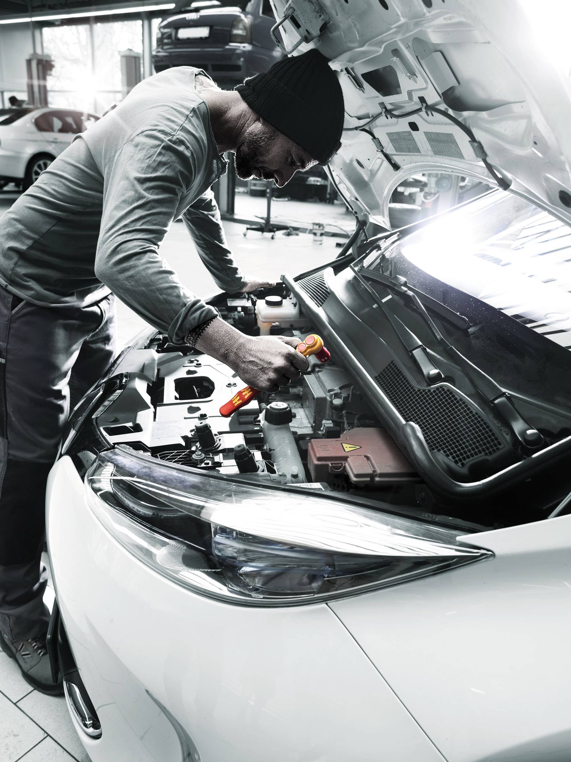 Ein Mann repariert einen Motor in einer Autowerkstatt. Er trägt eine Mütze und hält ein Werkzeug. Ein anderes Auto ist im Hintergrund.