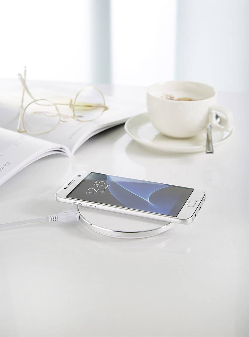 Ein Smartphone lädt kabellos auf einer Ladestation. Daneben liegen eine Brille, eine geöffnete Zeitschrift und eine Tasse Kaffee.