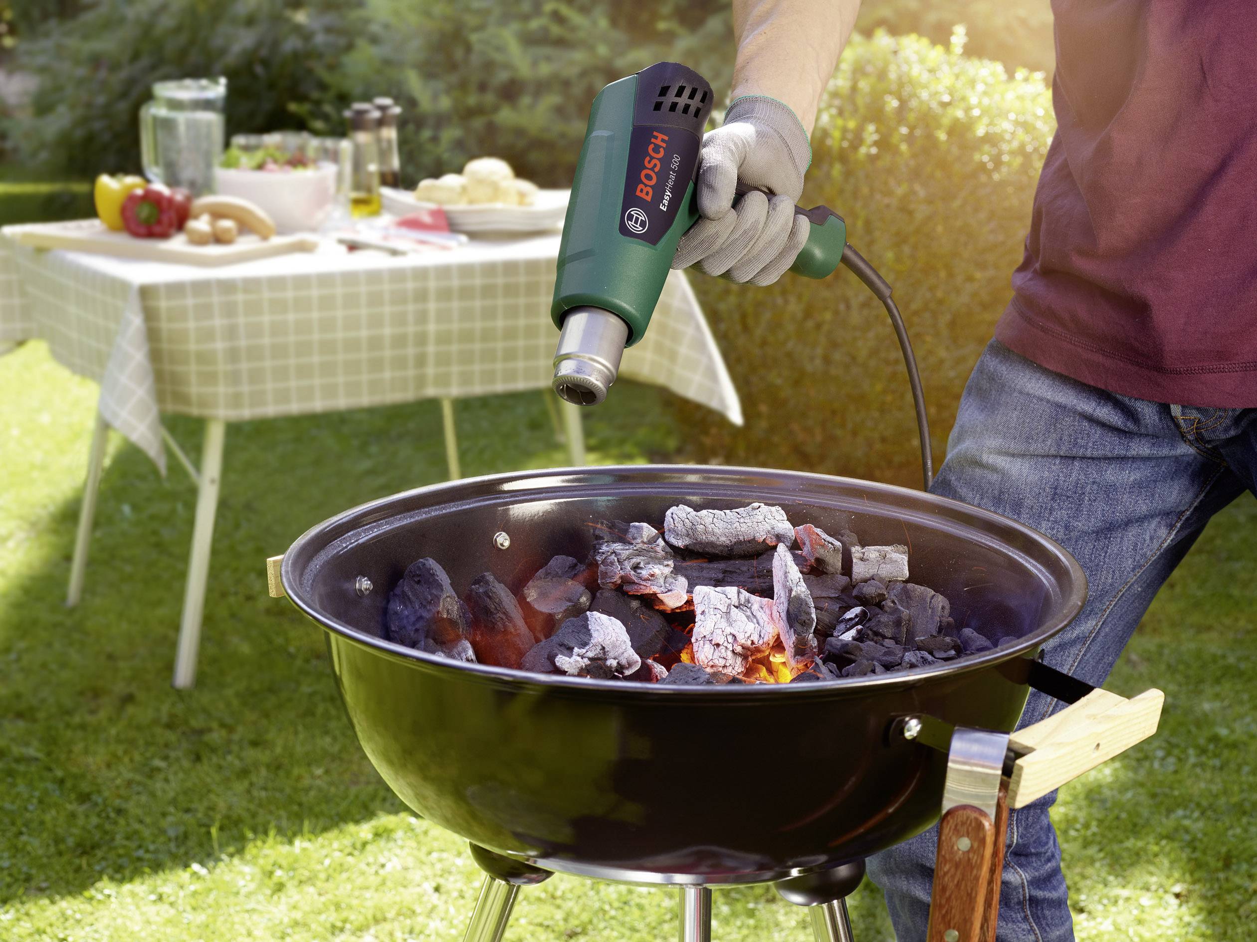Person erhitzt Grillkohle mit einem Heißluftgebläse im Garten. Im Hintergrund steht ein Tisch mit Essen und Getränken.