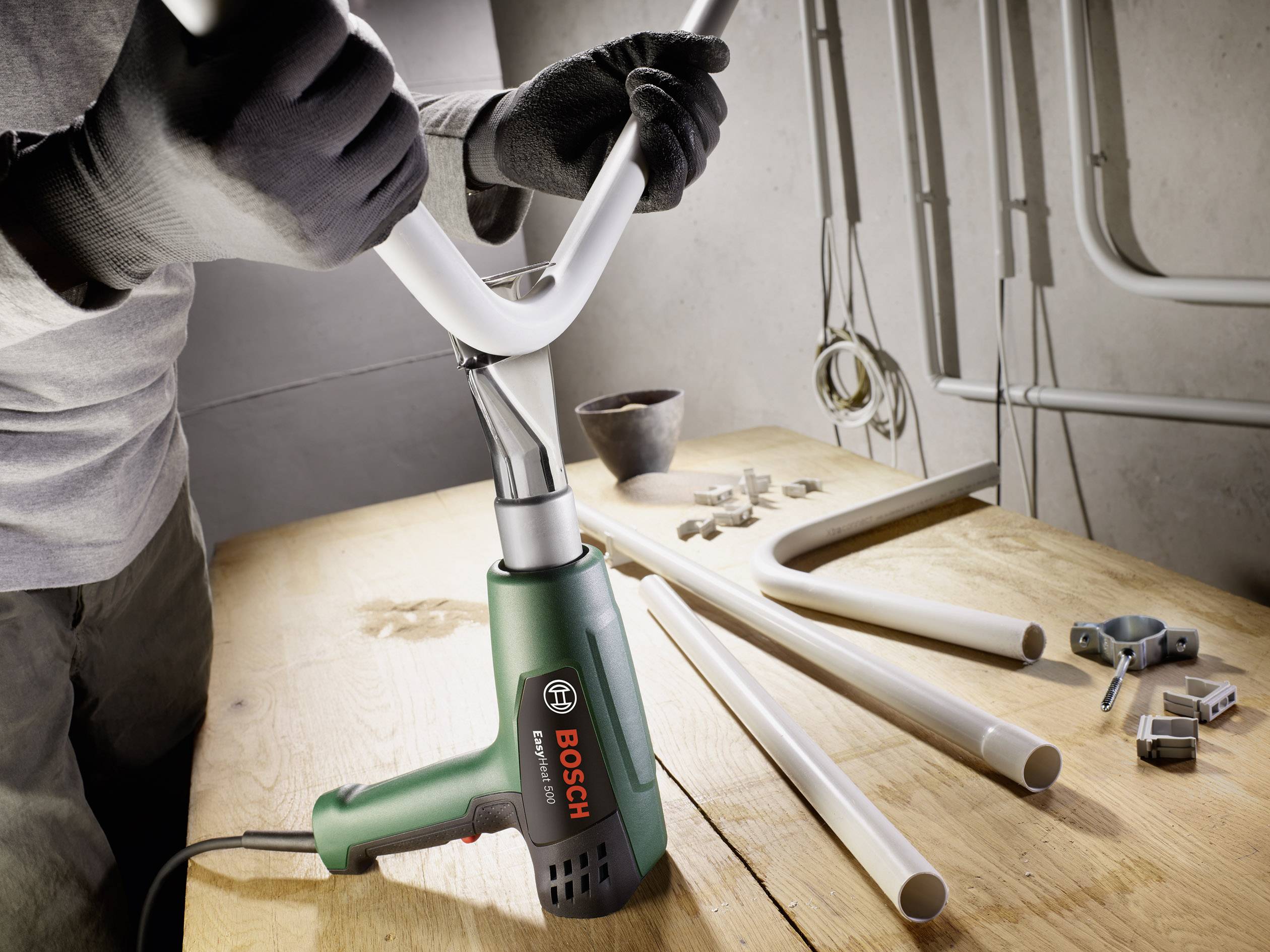 Eine Person biegt mit einem Bosch-Heißluftgebläse ein weißes Rohr auf einem Holztisch, umgeben von weiteren Rohren und Zubehör.