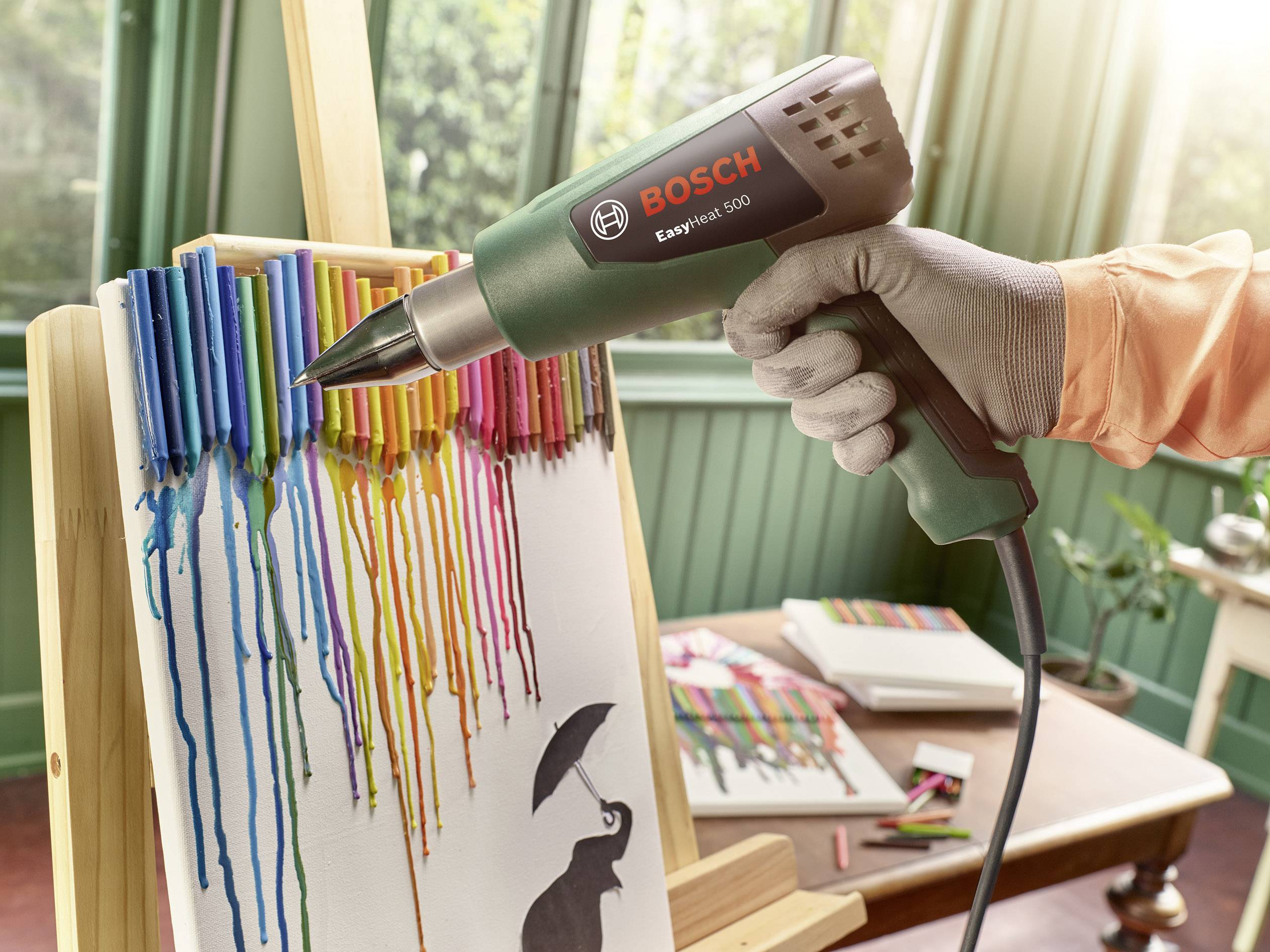 Ein Föhn schmilzt bunte Wachsmalstifte, die auf einer Leinwand befestigt sind. Die geschmolzenen Farben laufen tropfenförmig nach unten.