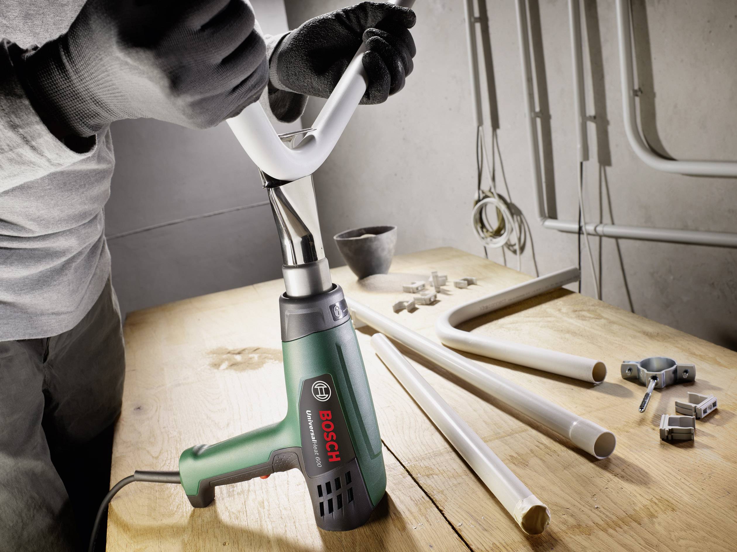Eine Person arbeitet mit einem Heißluftgebläse von Bosch an einem weißen Rohr auf einem Holztisch. Im Hintergrund sind weitere Werkzeuge und Rohre zu sehen.