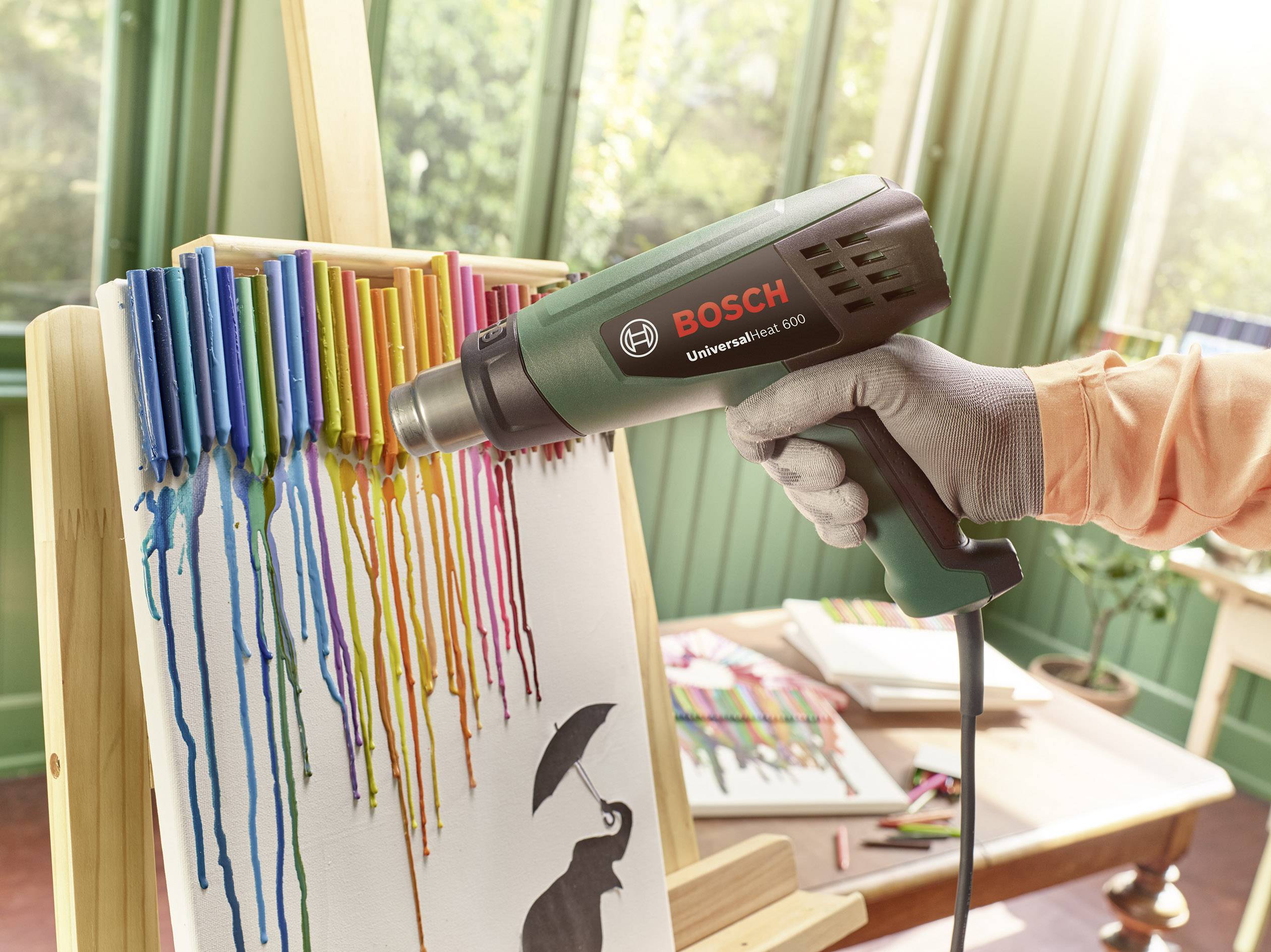 Eine Person verwendet ein Heißluftgebläse, um bunte Wachsmalstifte auf einer Leinwand zum Schmelzen zu bringen. Im Hintergrund ein grüner Raum.