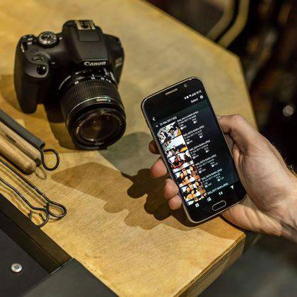 Eine Kamera steht auf einem Tisch neben einem Smartphone, das in einer Hand gehalten wird. Auf dem Handybildschirm sind Fotos zu sehen.