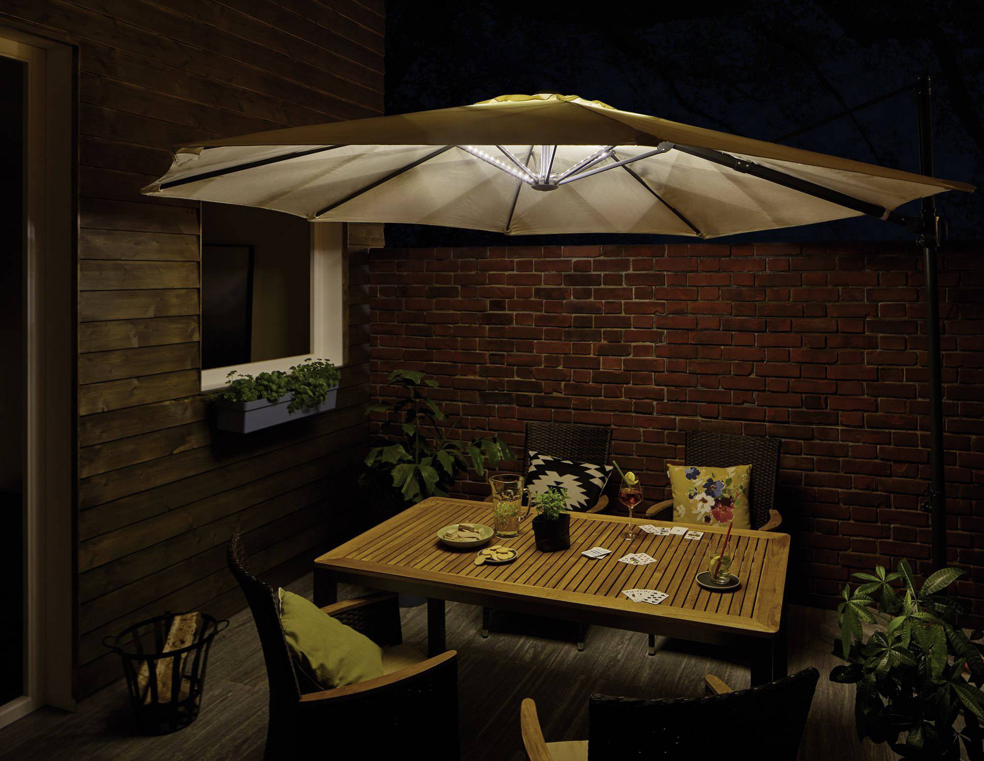 Ein beleuchteter Terrassentisch bei Nacht, umgeben von Pflanzen und Stühlen mit Kissen. Ein großer Sonnenschirm spendet Licht.