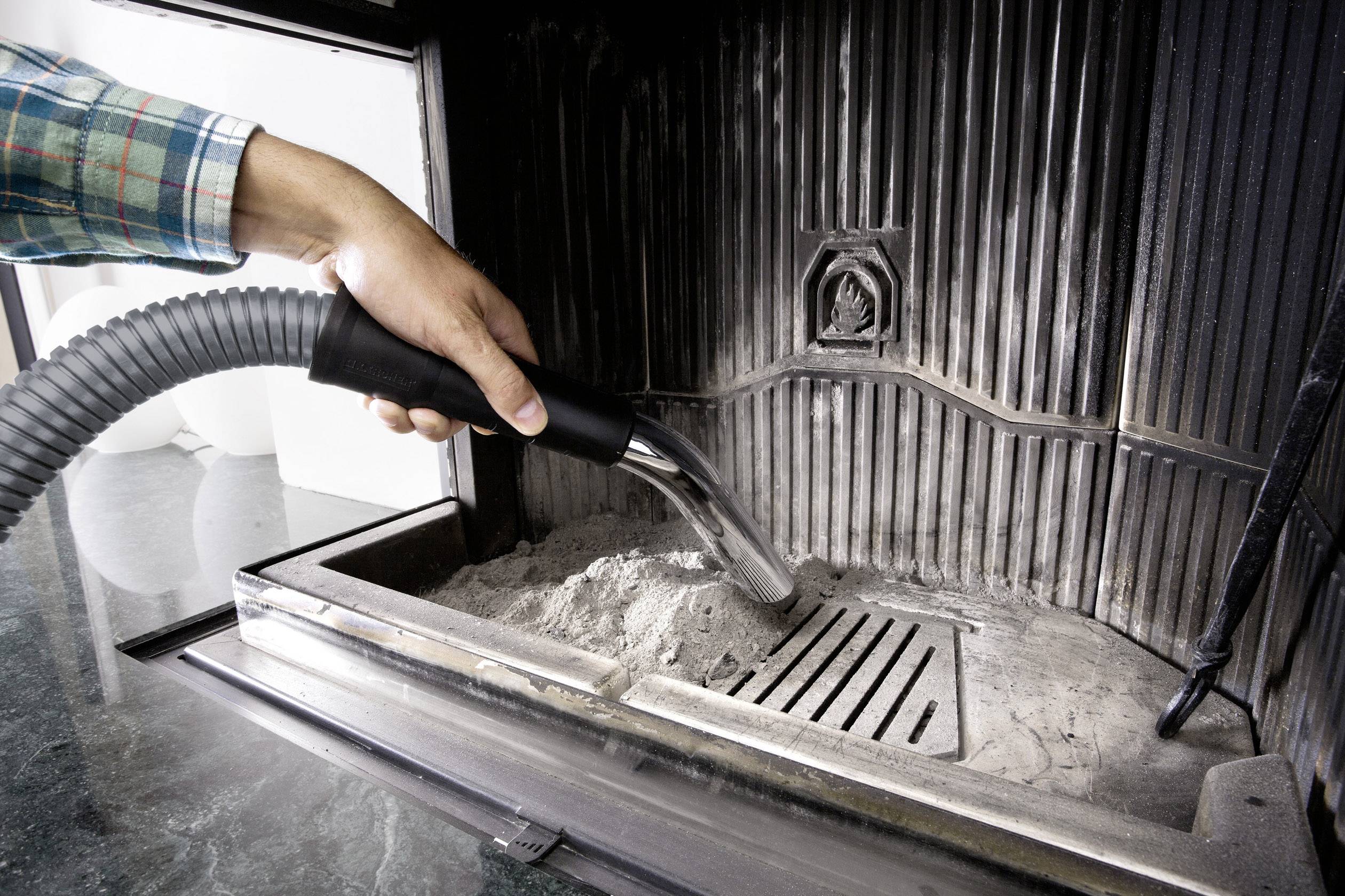 Eine Person reinigt die Asche aus einem Kamin mit einem Staubsauger. Der Kamin ist aus Metall, und das Bild zeigt den Reinigungsprozess.