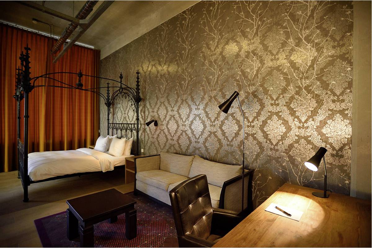 Ein stilvolles Hotelzimmer mit Himmelbett, beigem Sofa, Holztisch und Lampe. Die Wände sind mit dekorativen Mustern verziert.