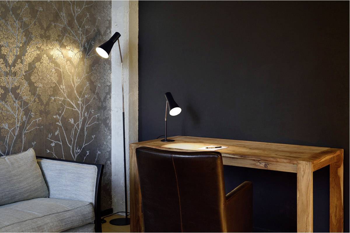 Ein minimalistischer Arbeitsbereich mit einem Holztisch, einer schwarzen Lampe und einem Ledersessel vor einer dunklen Wand. Links steht eine Couch.