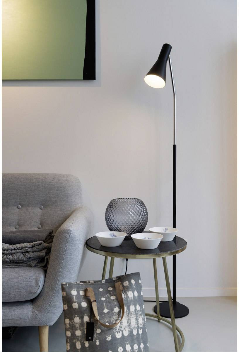 Ein gemütliches Wohnzimmer mit grauem Sessel, Stehlampe, rundem Tisch mit Schalen und Vase. Im Vordergrund eine gemusterte Tasche.