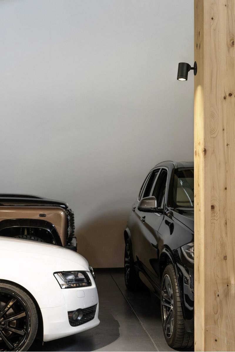 Zwei parkende Autos in einer Garage, teilweise verborgen hinter einer Holzwand. Links ein weißes Fahrzeug, rechts ein schwarzes SUV.