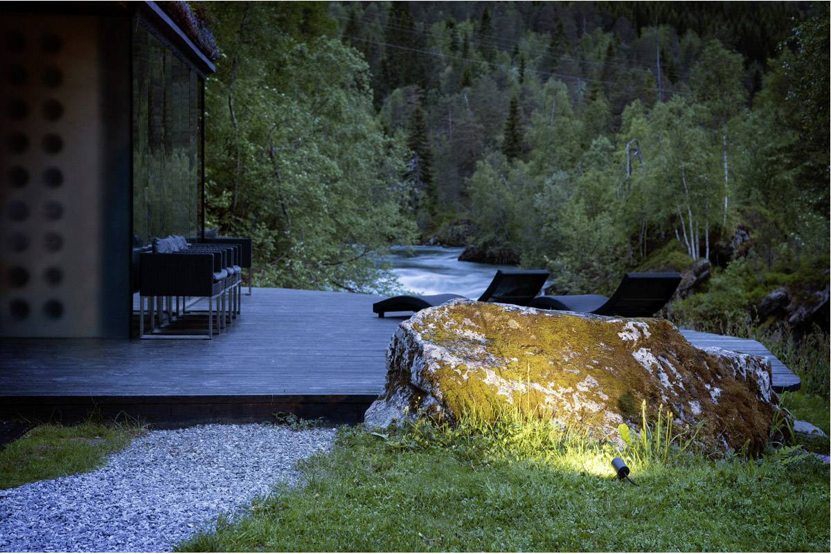 Eine Terrasse mit Liegestühlen und Tisch grenzt an einen Fluss in bewaldeter Umgebung; am Boden beleuchtet ein Strahler einen großen Stein.