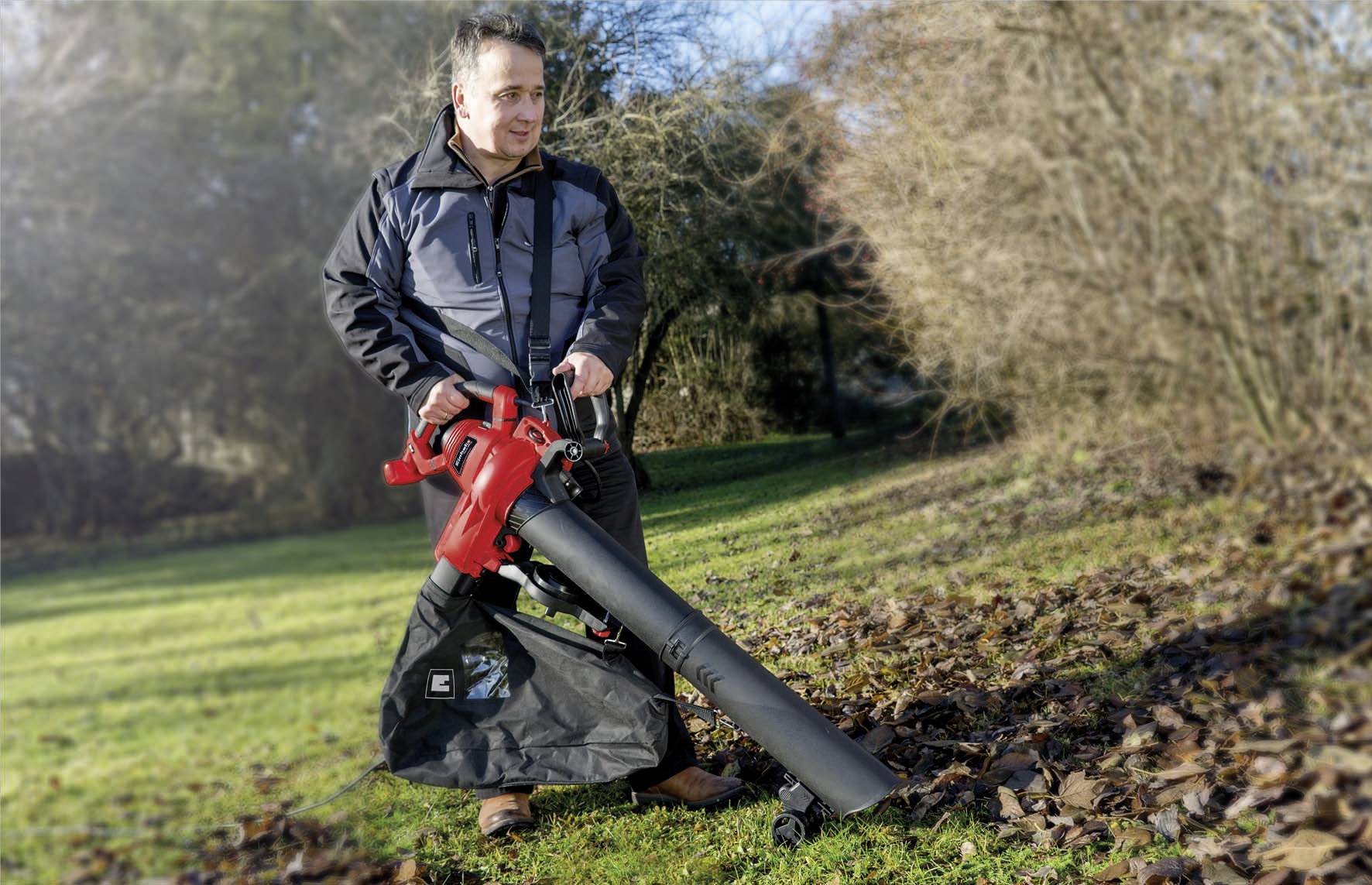 Ein Mann benutzt einen roten Laubsauger auf einer Wiese, um Laub zu entfernen. Er trägt eine Jacke und steht neben Bäumen im Hintergrund.
