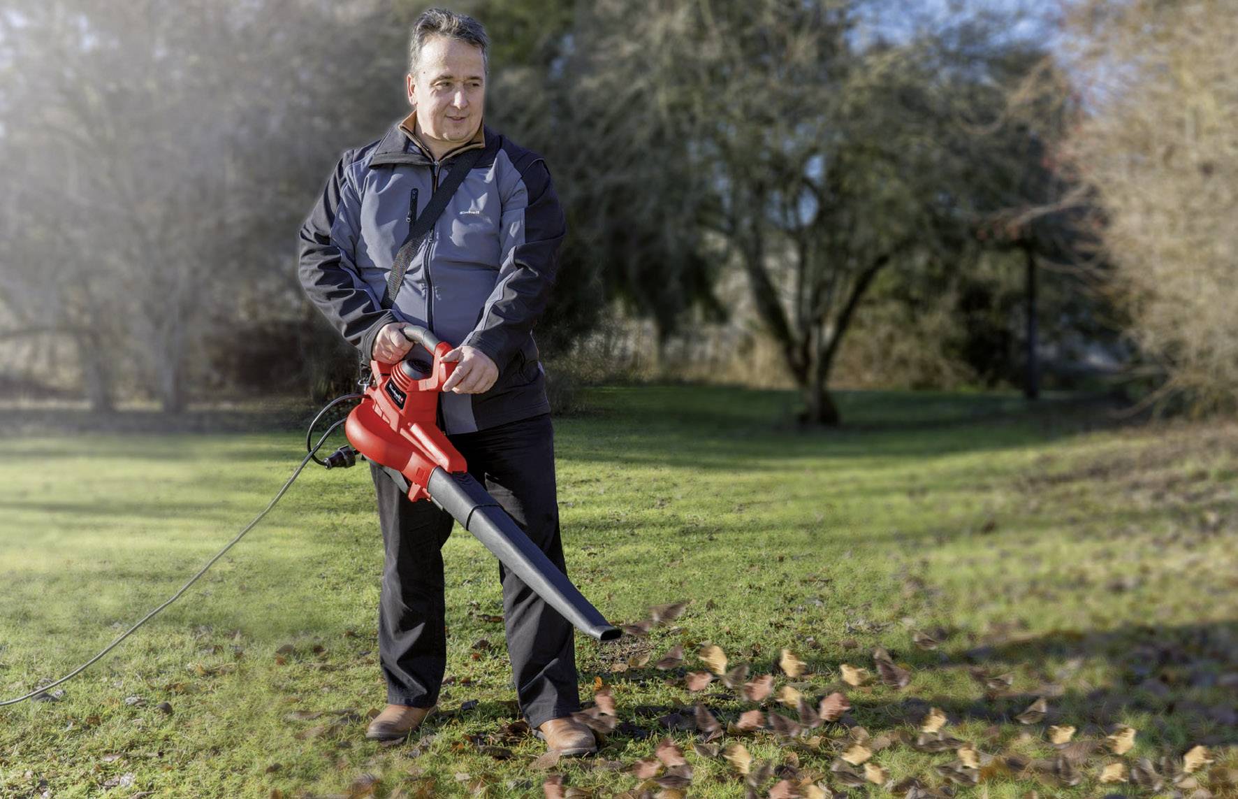 Ein Mann mit einer dunklen Jacke benutzt einen roten Laubbläser, um Herbstlaub auf einer grünen Rasenfläche zu entfernen.