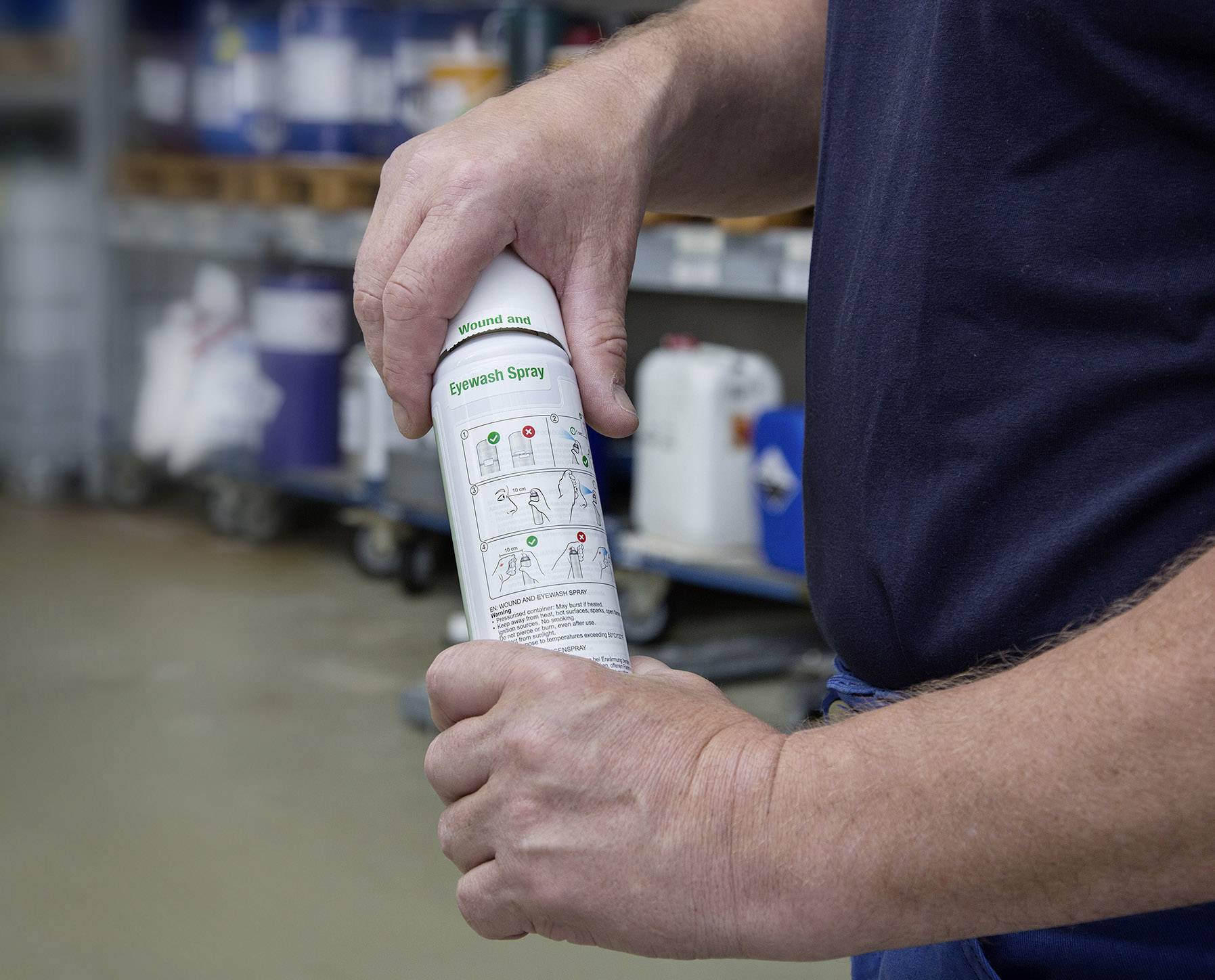 Eine Person hält ein Augenspül-Spray, bereit zur Anwendung. Im Hintergrund sind Regale mit verschiedenen Behältern zu sehen.