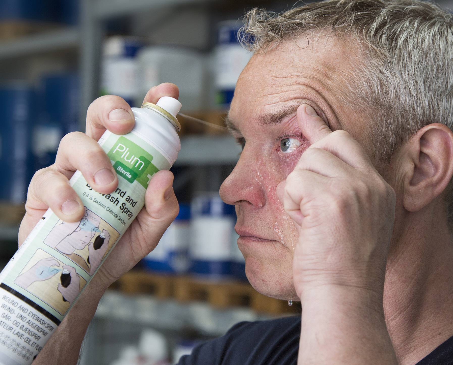 Mann sprüht Augenspülung in sein Auge zur Reinigung. Flüssigkeit tropft von seinem Gesicht. Im Hintergrund Regale mit Produkten.