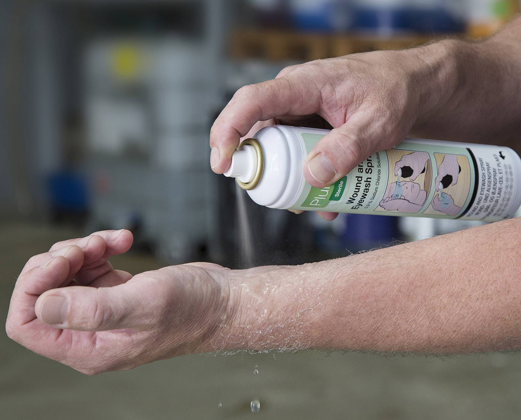 Eine Person sprüht einen Erste-Hilfe-Spray auf den Unterarm, um eine Verletzung oder Irritation zu behandeln. Im Hintergrund ist eine verschwommene Werkstatt zu sehen.