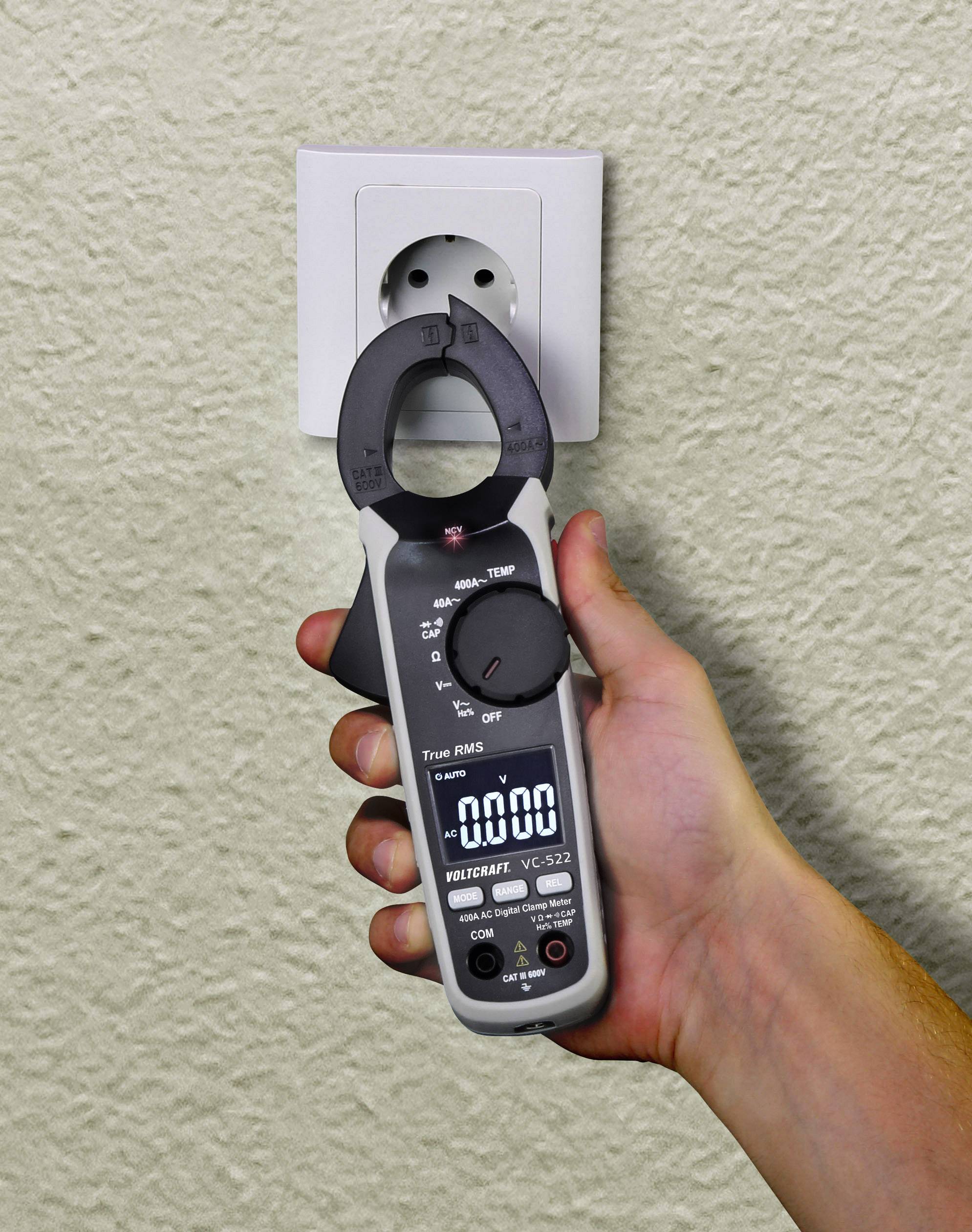 Eine Hand hält ein Spannungsmessgerät vor einer Steckdose. Das Messgerät zeigt 