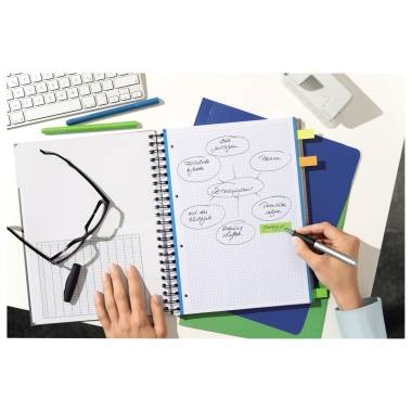 Eine Person schreibt mit einem Stift in ein geöffnetes Notizbuch auf einem Schreibtisch, umgeben von Brille, Tastatur und Büromaterial.