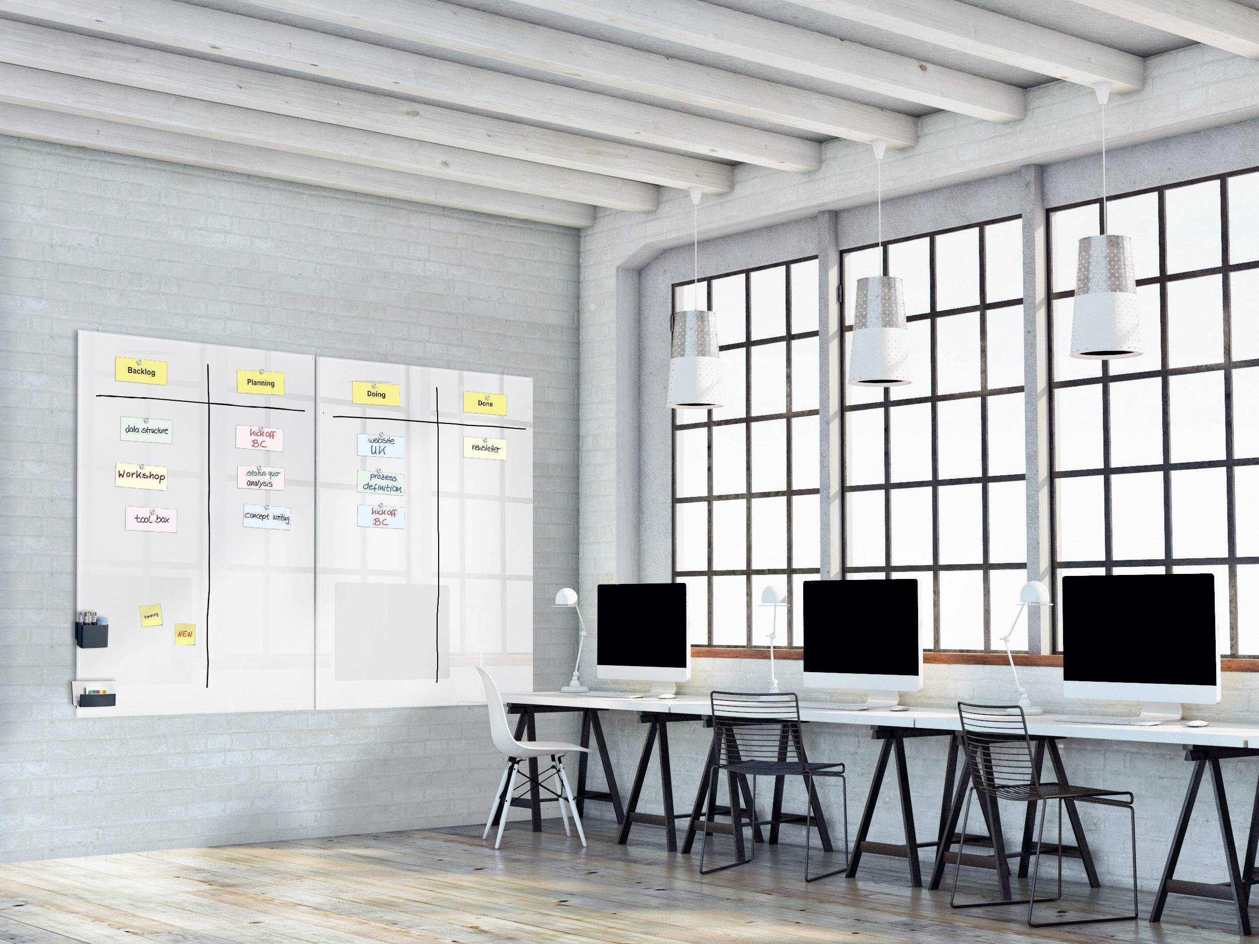 Helles Büro mit fünf Computern an einem Tisch. An der Wand ein Whiteboard mit Notizen, das einen Arbeitsprozess zeigt.