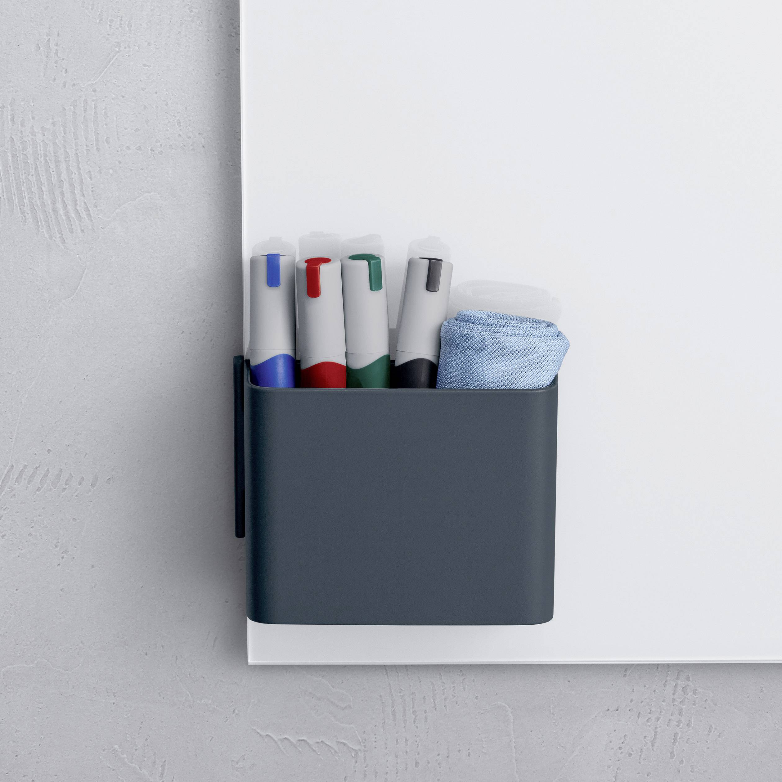 Whiteboard mit Halterung für vier farbige Stifte und ein Löschpad, an einer grauen Wand befestigt.