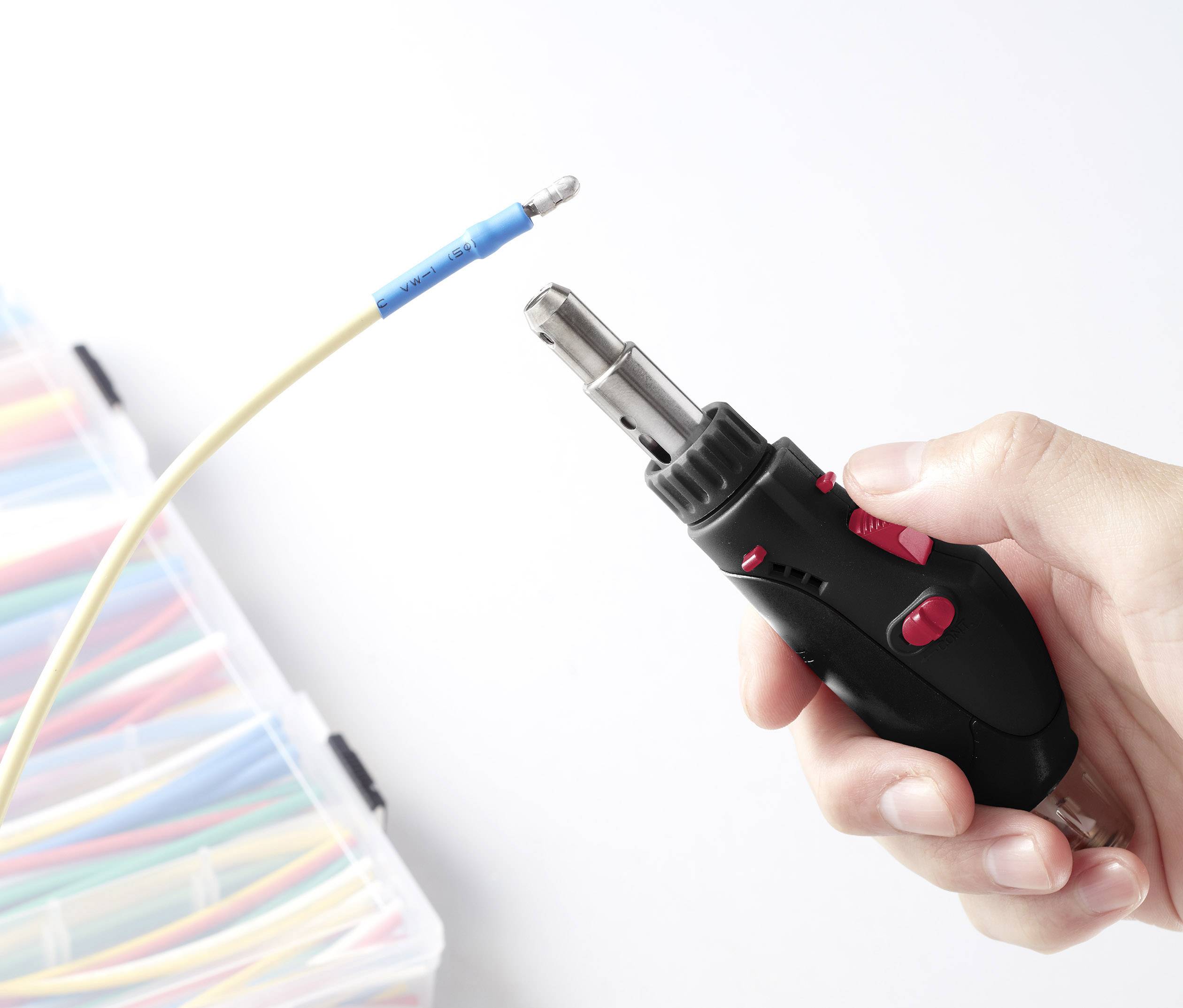 Eine Hand hält einen Gasbrenner, der auf ein gelbes Kabel mit blauer Kunststoffhülle gerichtet ist; im Hintergrund ist ein Organizer mit bunten Kabeln.