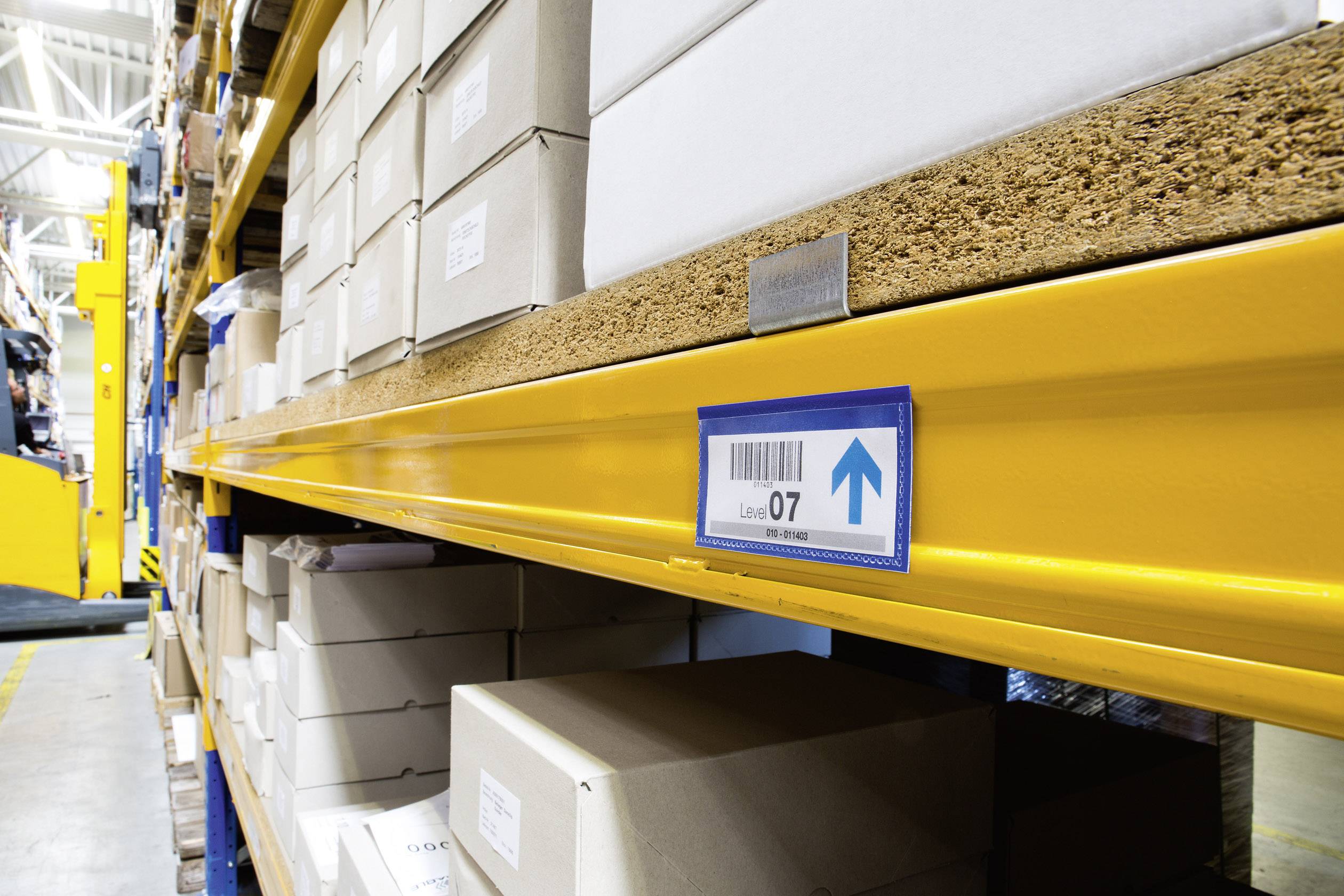 Ein Metallregal in einem Lagerhaus ist mit Kartons gefüllt. Ein blauer Pfeil auf einem Schild zeigt aufwärts. Im Hintergrund ein Gabelstapler.