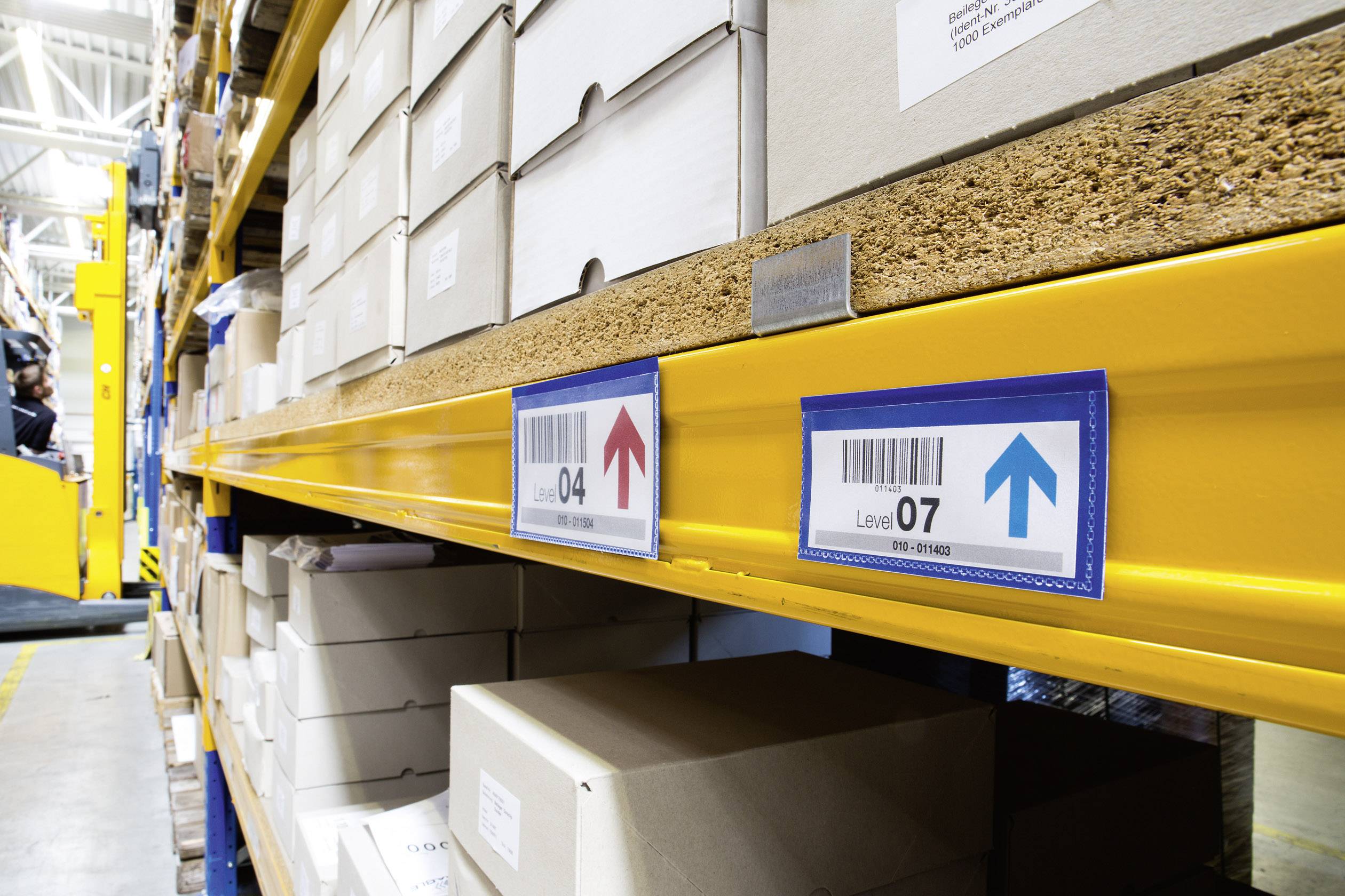 Regale mit Kartons in einem Lager. Etiketten mit Barcodes und Pfeilen zeigen Lagerorte an. Ein Gabelstapler ist im Hintergrund zu sehen.
