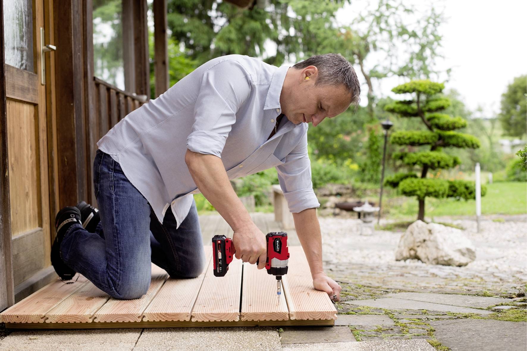 Ein Mann kniet auf einer Holzterrasse und schraubt mit einem Akkuschrauber Bretter fest. Im Hintergrund ein gepflegter Garten.