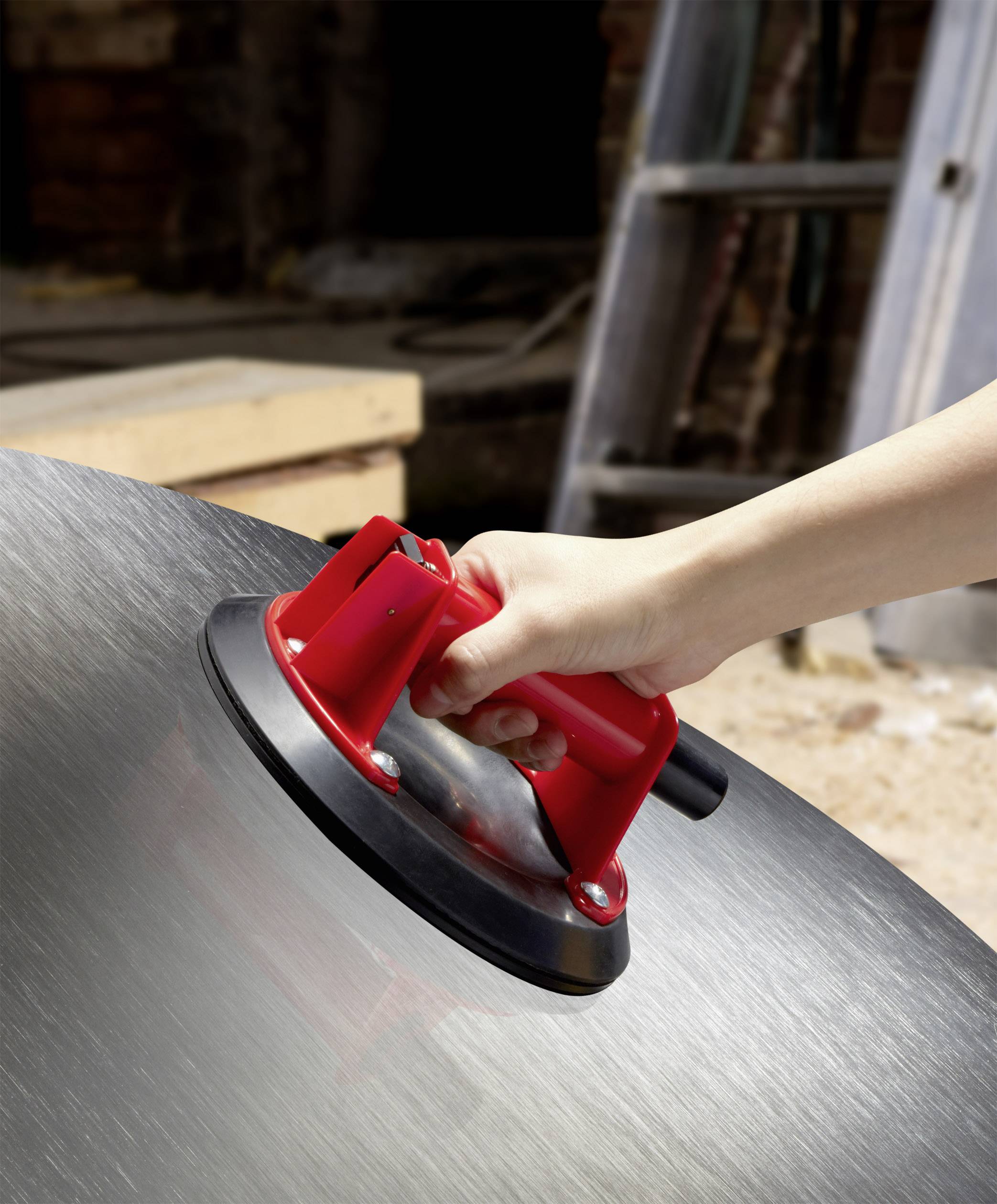 Eine Hand hält einen roten Saugheber an einer großen, glänzenden Metalloberfläche, offenbar zum Transport schwerer oder glatter Gegenstände.