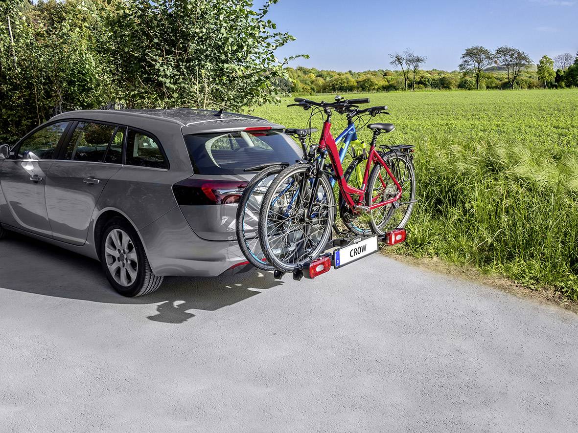 Auto mit Fahrradträger und drei Fahrrädern, geparkt auf einer ländlichen Straße. Im Hintergrund grüne Felder und Bäume.
