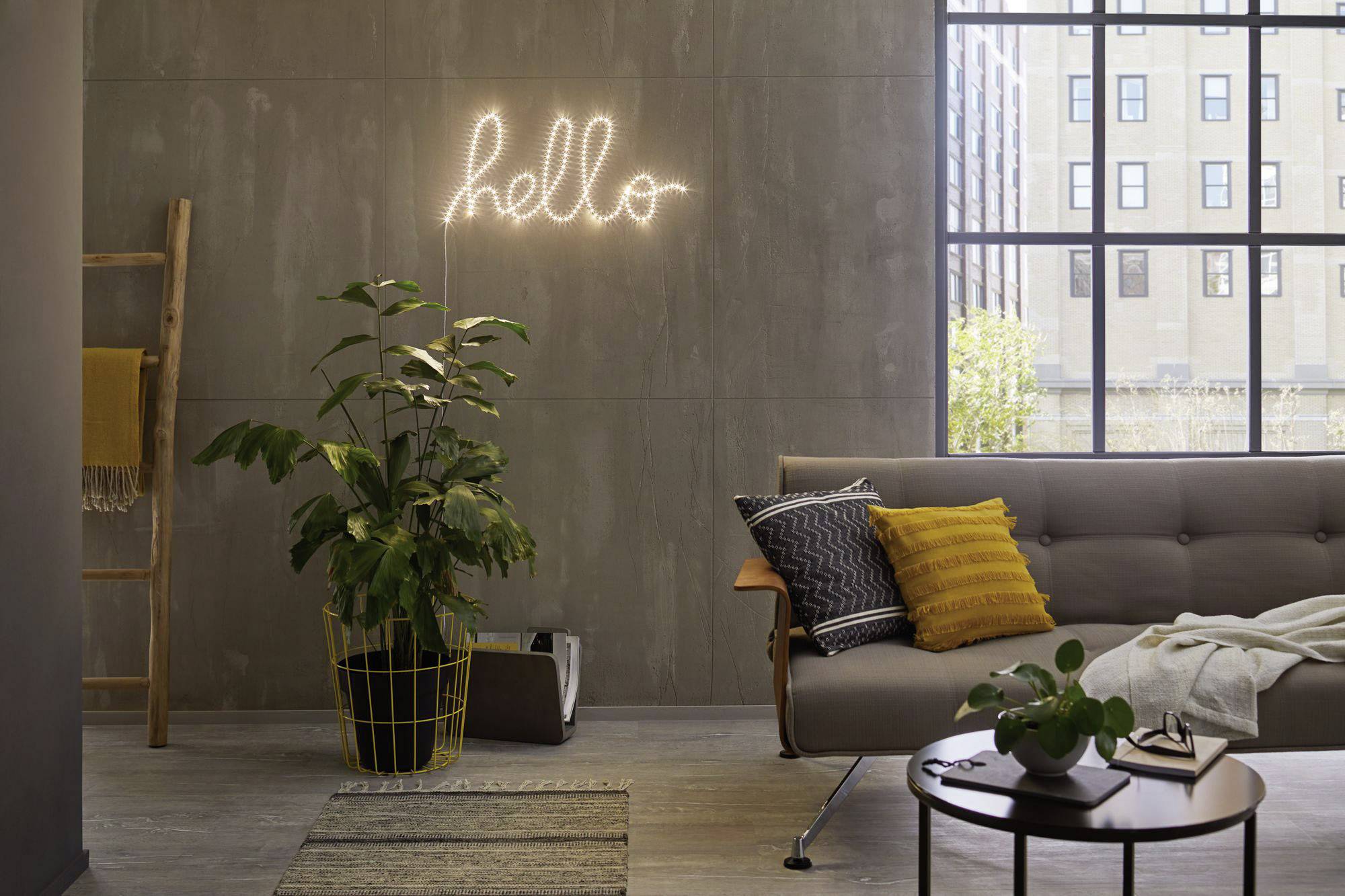 Ein Wohnzimmer mit grauem Sofa, Kissen, Pflanze im Korb und Teppich. An der Wand hängt ein leuchtendes 'hello'-Schild. Großer Fensterblick.