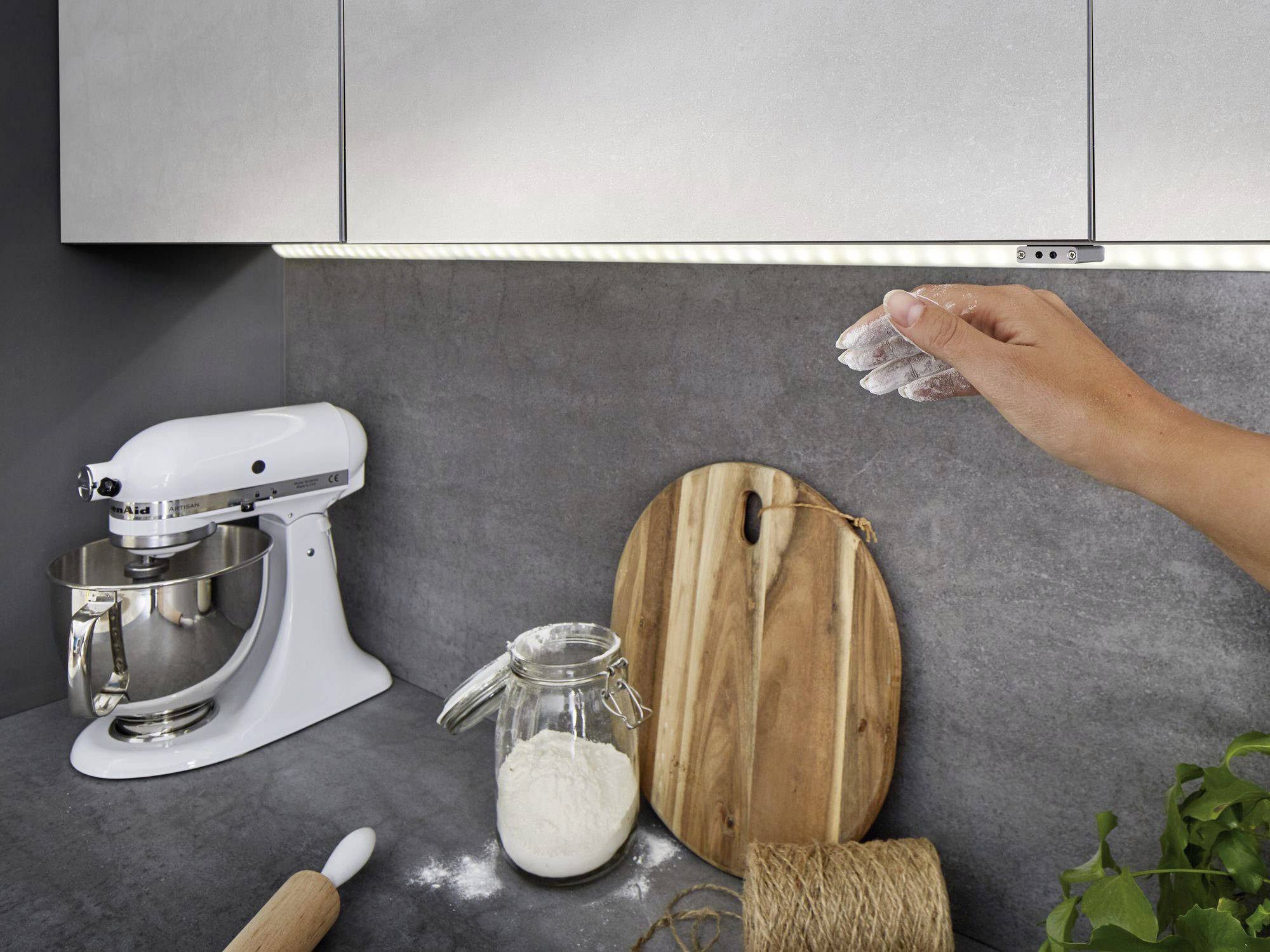 Hand streut Mehl über die Arbeitsfläche in moderner Küche. Ein Mixer, ein Glas Mehl und Holzbretter sind sichtbar.