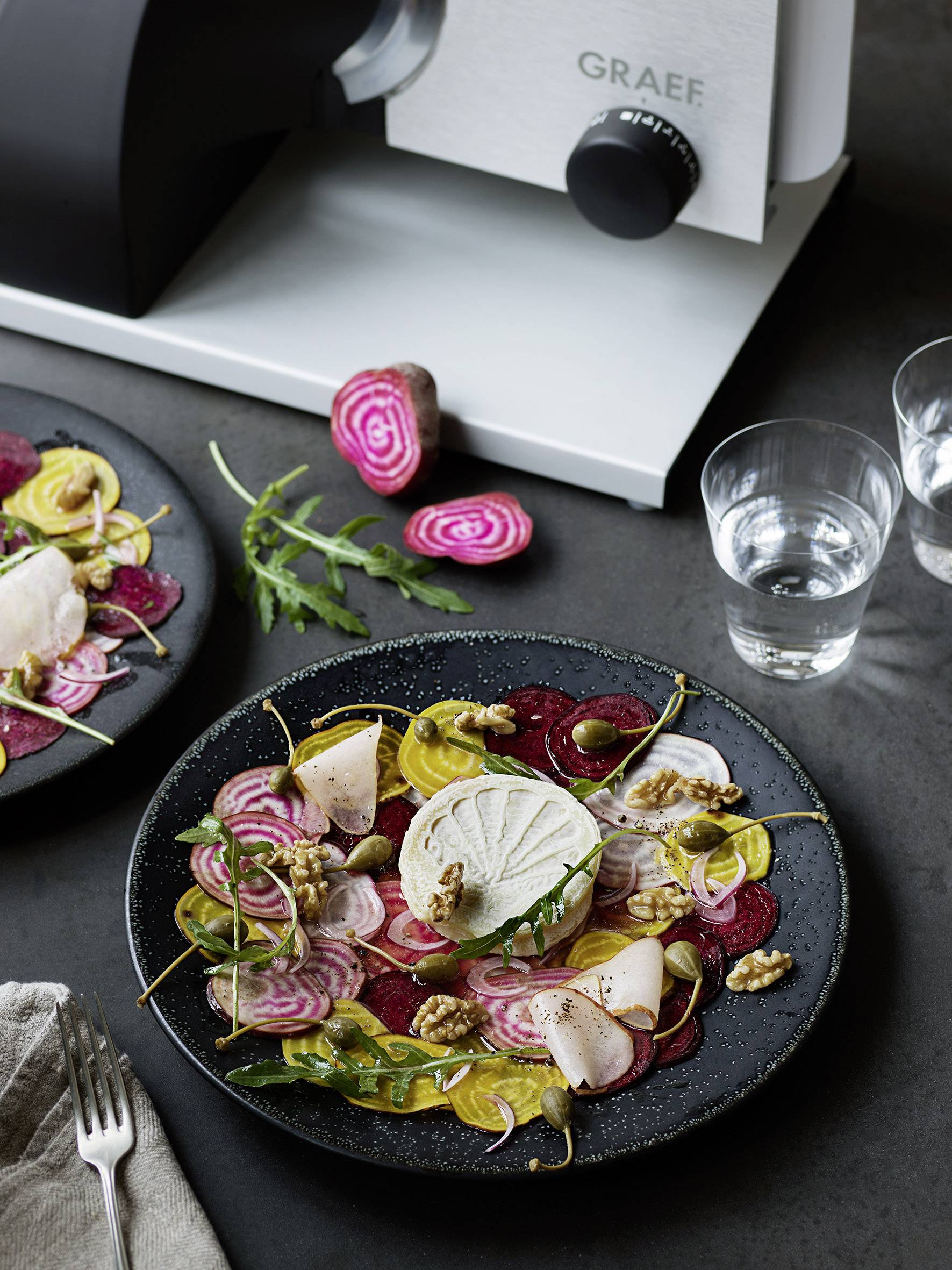 Ein Teller mit bunten Rüben, Käse und Walnüssen, umgeben von Wassergläsern und einem Gemüseschneider im Hintergrund.