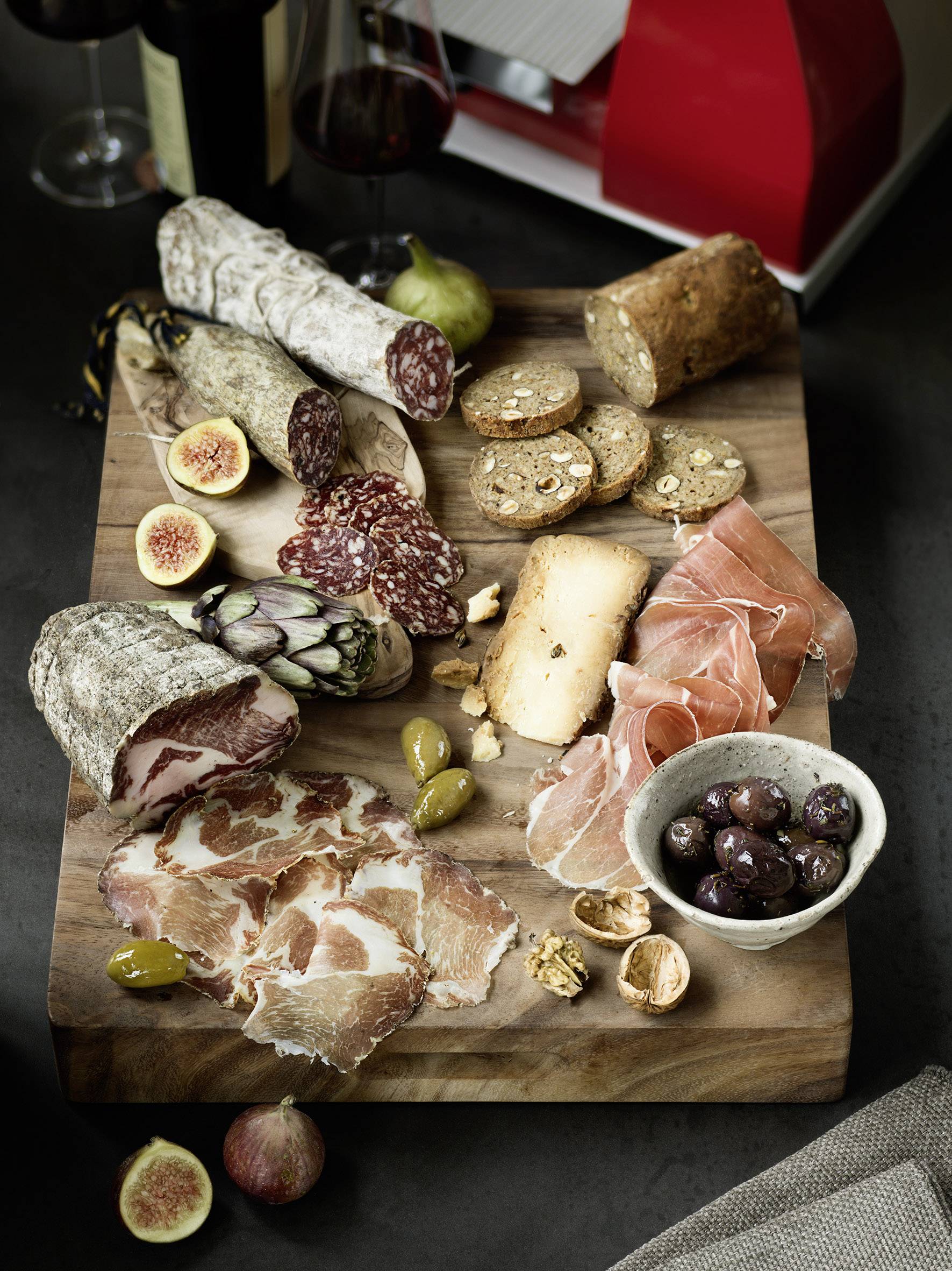 Auf einem Holzbrett liegt eine Auswahl an Aufschnitt, Würsten, Oliven und Feigen, umgeben von Brot und Crackern, mit Wein im Hintergrund.