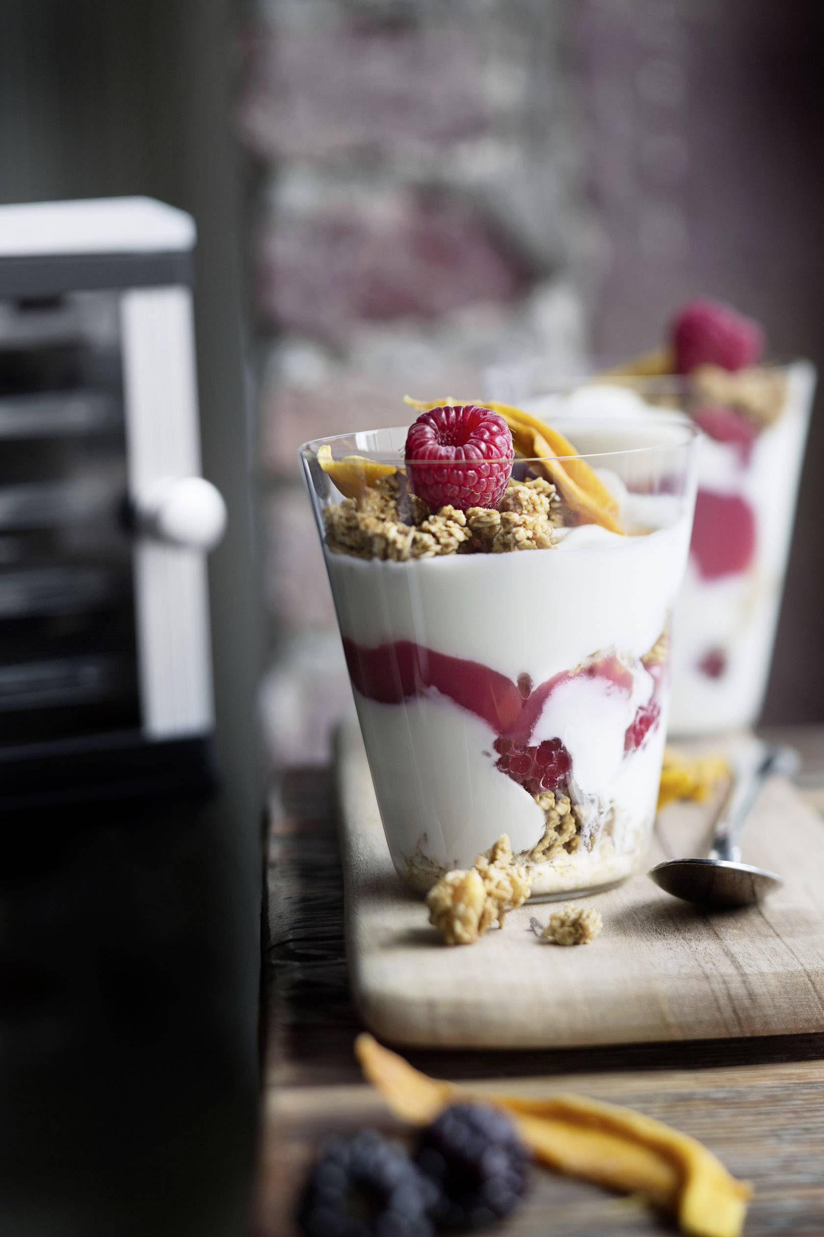 Ein Glas Schichtdessert mit Joghurt, Himbeeren, Müsli und getrockneten Mango-Streifen, auf einem Holzbrett neben einem Löffel.