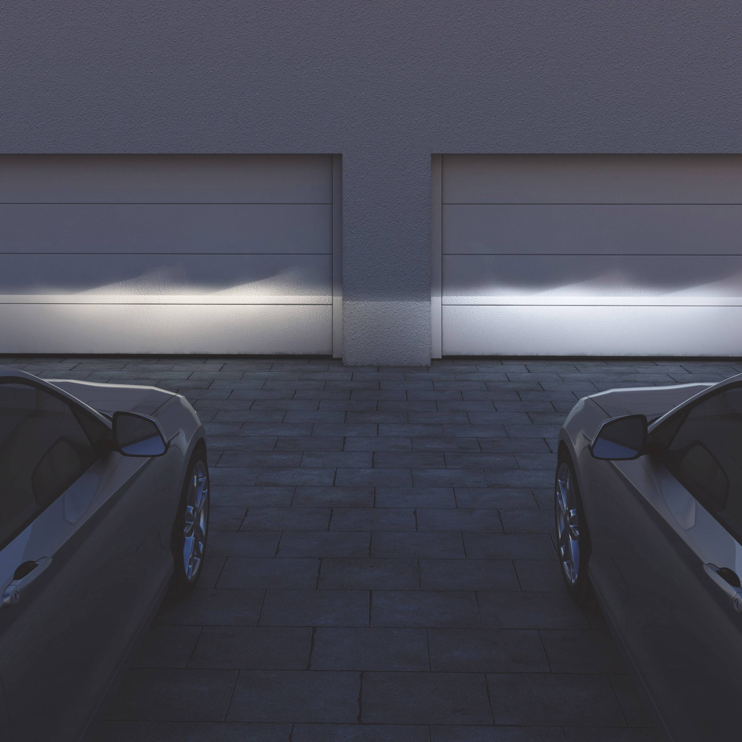 Zwei Autos stehen vor geschlossenen Garagentoren, beleuchtet von hellen Lichtern. Die Szene hat eine ruhige, abendliche Atmosphäre.