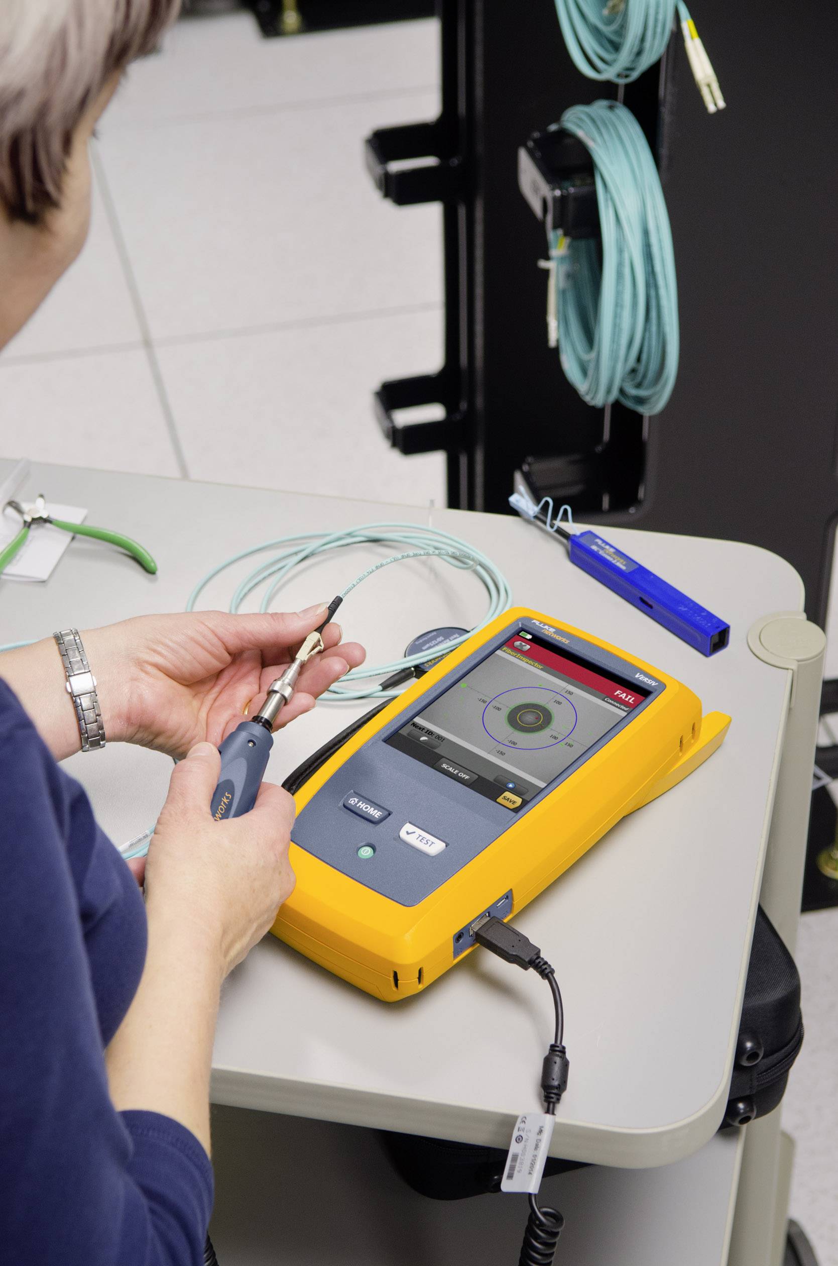Eine Person testet Glasfaserkabel mit einem Handmessgerät auf einem Tisch. Das Gerät zeigt Testresultate an. Kabel sind in der Nähe aufgewickelt.
