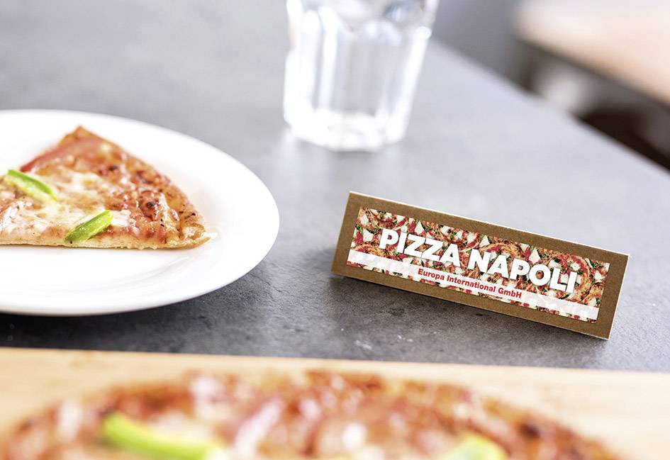 Ein Pizzastück auf einem Teller neben einem Schild mit der Aufschrift 'Pizza Napoli'. Im Hintergrund steht ein Glas Wasser.