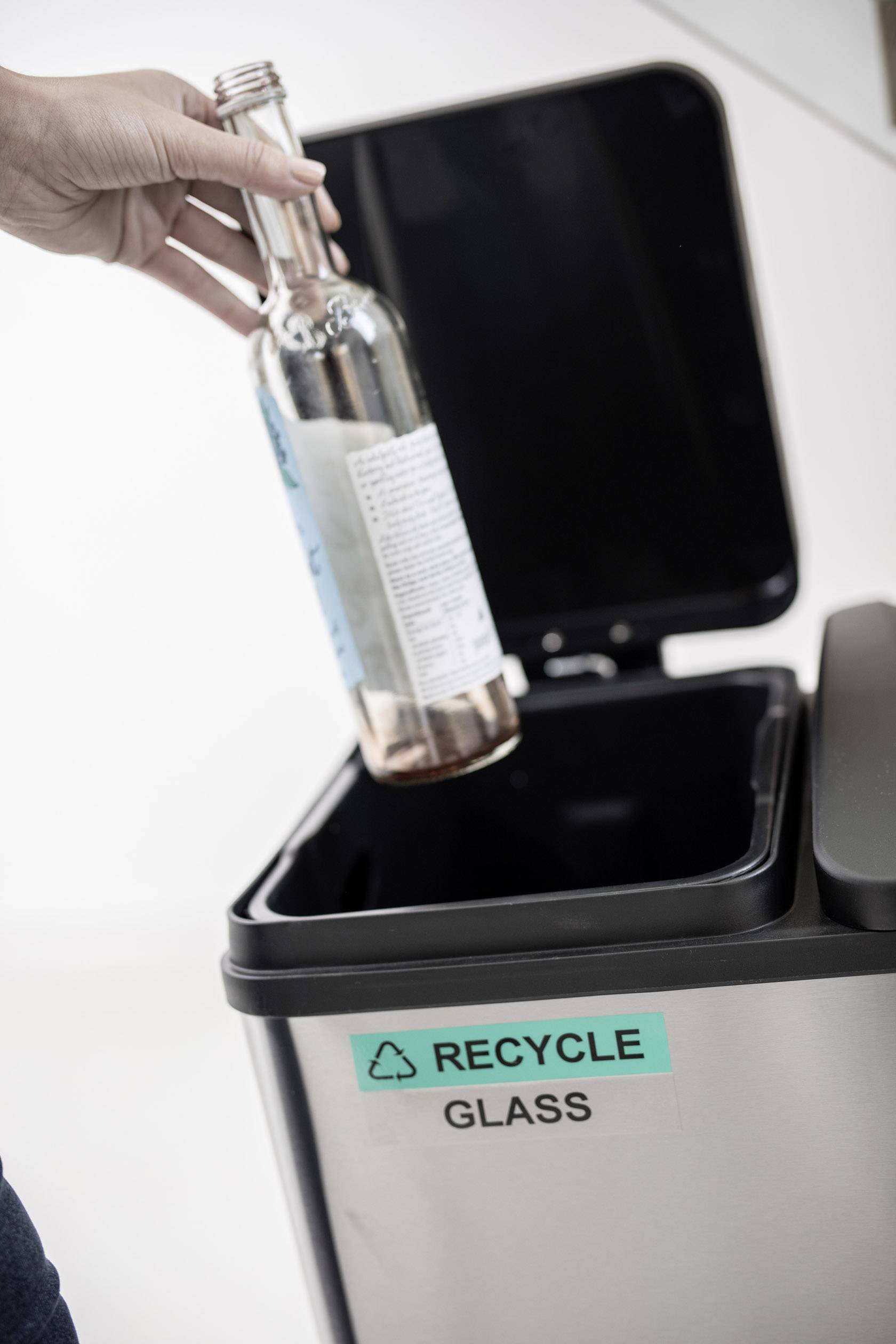 Eine Person wirft eine leere Glasflasche in einen Mülleimer mit der Aufschrift 'Recycle Glass'.