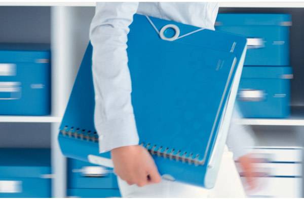 Person trägt einen großen, blauen Aktenordner; Hintergrund mit Regal voller blauer Kisten.