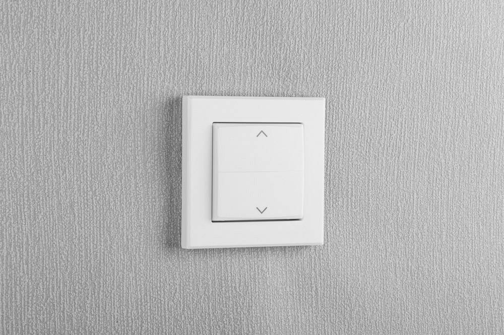 Ein weißer Lichtschalter mit zwei Tasten für Auf- und Abwärtsbewegung an einer grauen Wand montiert.