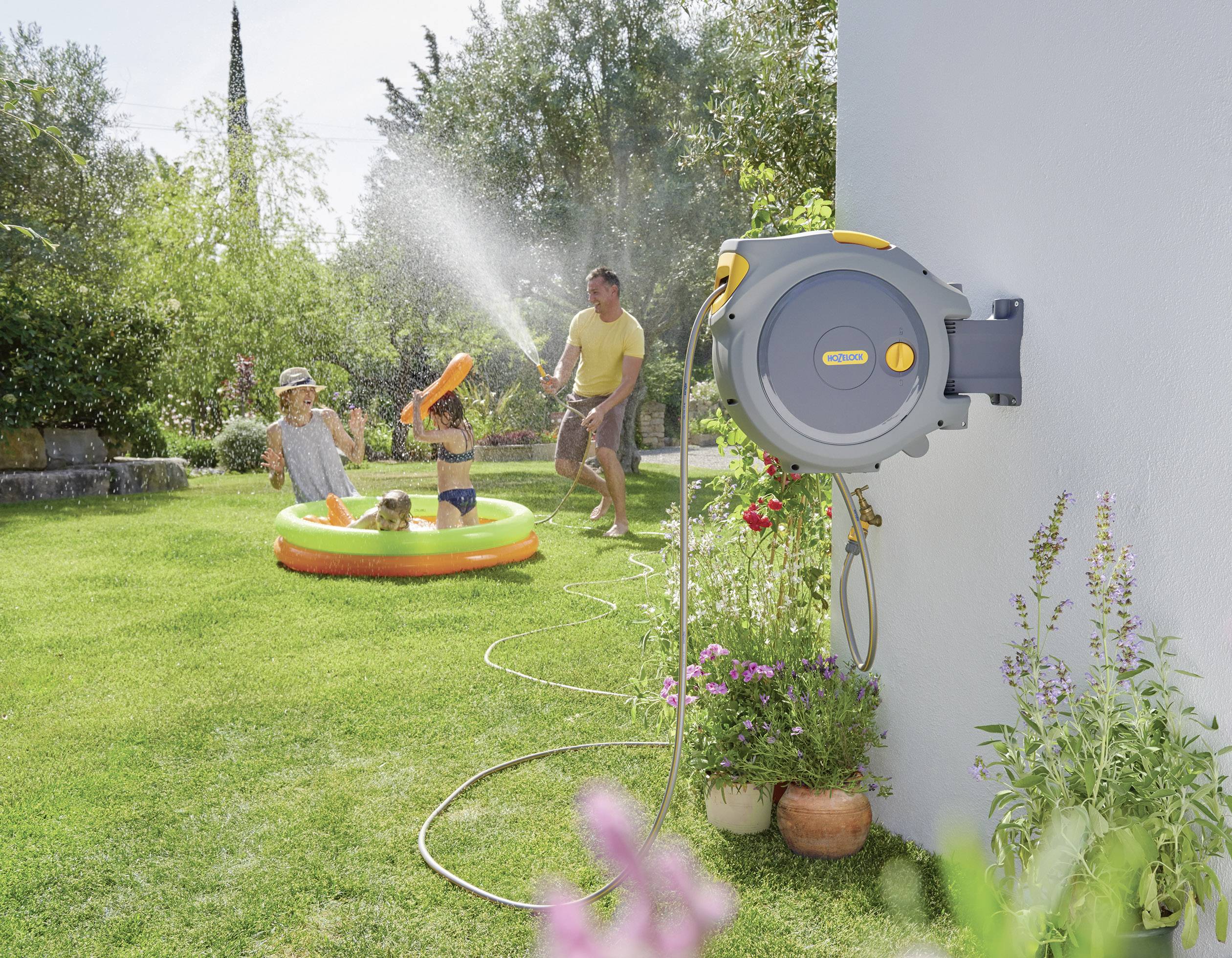 Eine Familie spielt in einem Garten: ein Erwachsener spritzt zwei Kinder mit Wasser aus einem Schlauch, während ein Hund im Planschbecken sitzt.