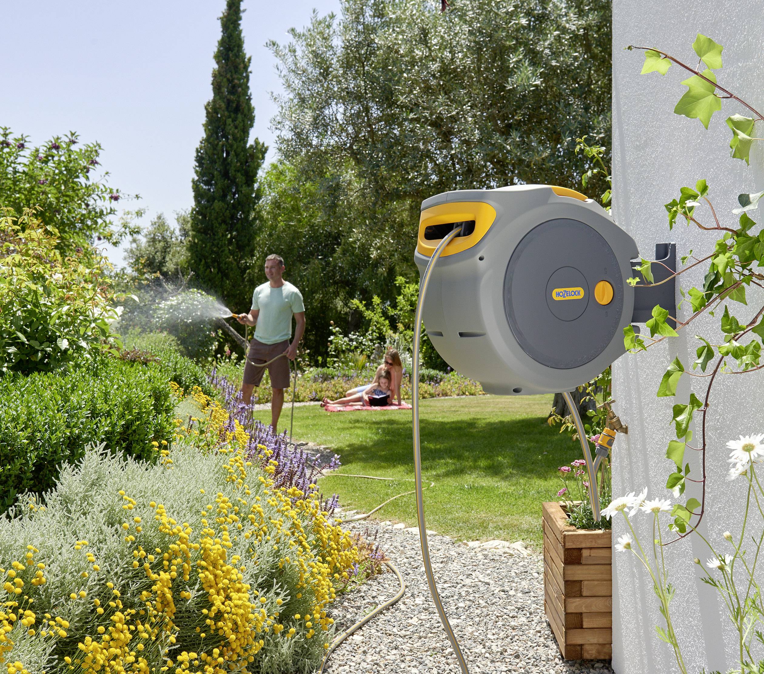 Ein Mann gießt einen Garten mit bunten Blumen, während ein Kind auf dem Rasen sitzt. Eine gelbe Schlauchtrommel ist an der Wand befestigt.