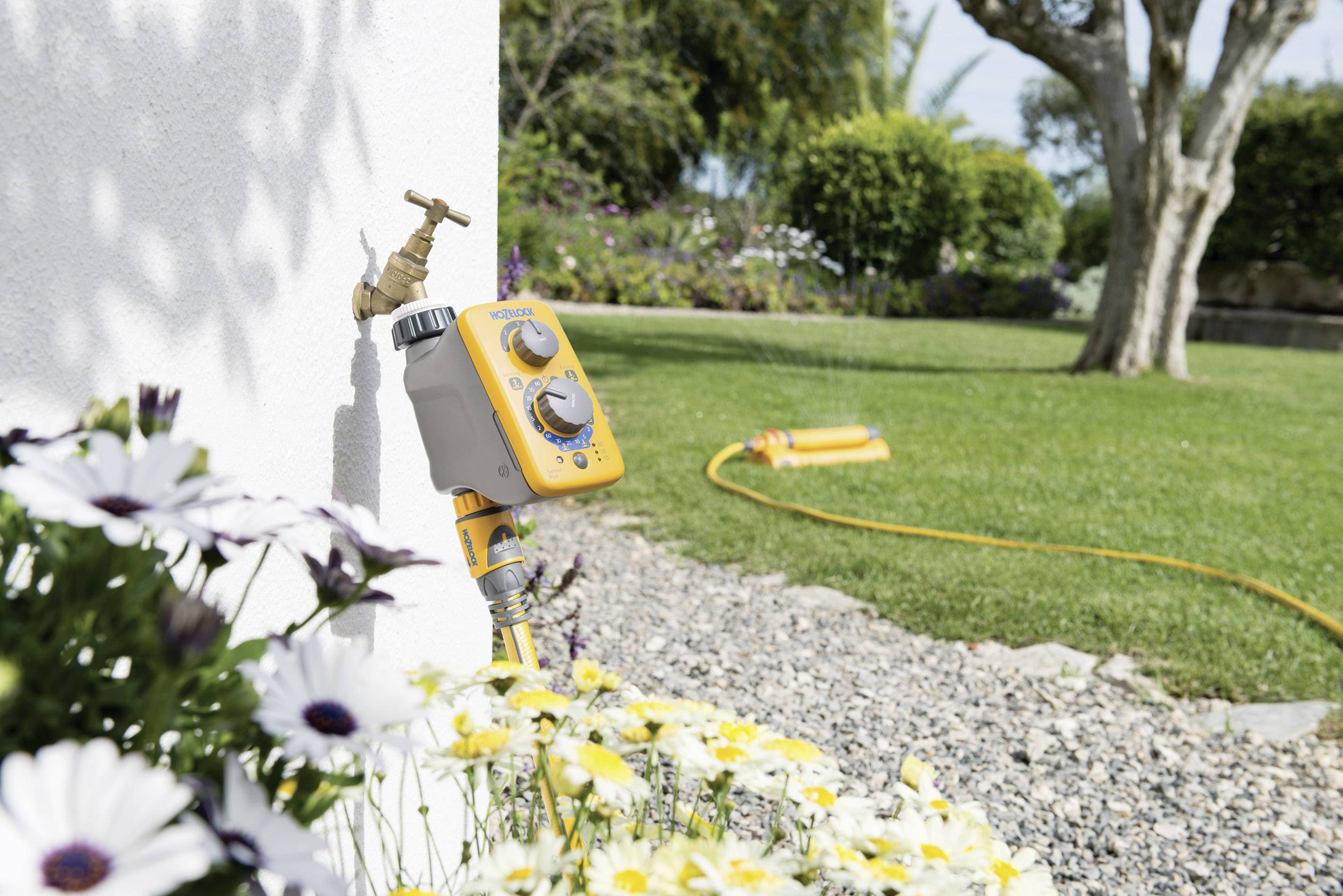 Ein automatischer Bewässerungs-Timer ist an einen Außenwasserhahn angeschlossen. Ein Gartenschlauch mit Sprinkler ist an den Timer angeschlossen.