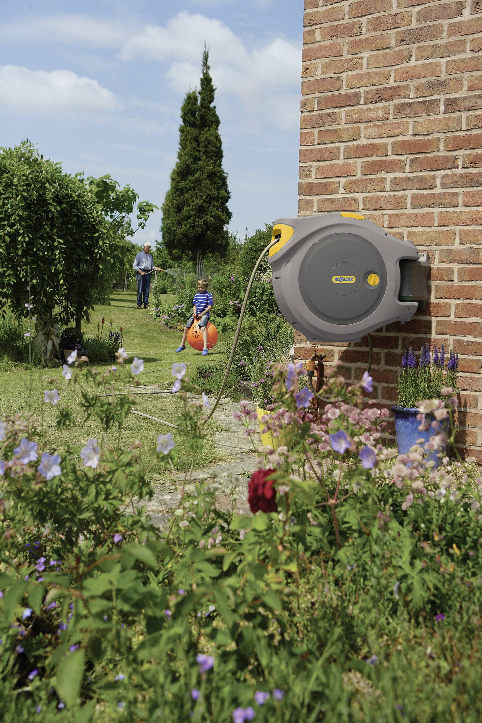 Eine automatische Schlauchtrommel ist an einer Backsteinmauer im blühenden Garten befestigt. Ein Kind spielt im Hintergrund.