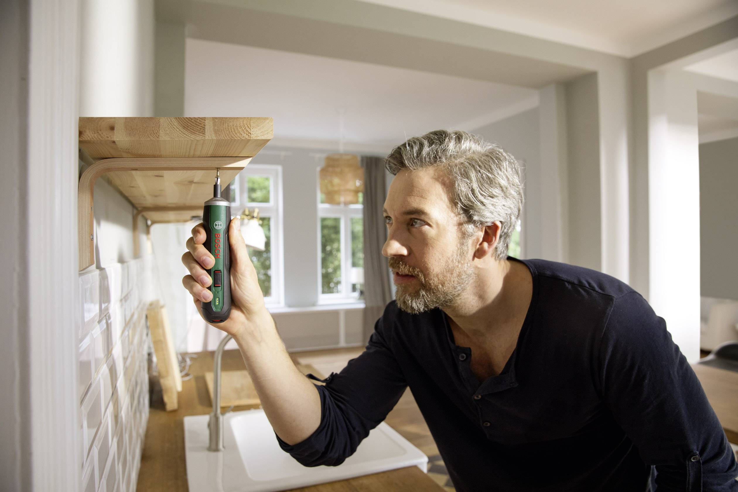 Mann montiert Holzregal an der Wand mit einem elektrischen Schraubendreher. Im Hintergrund ist ein helles Wohnzimmer sichtbar.