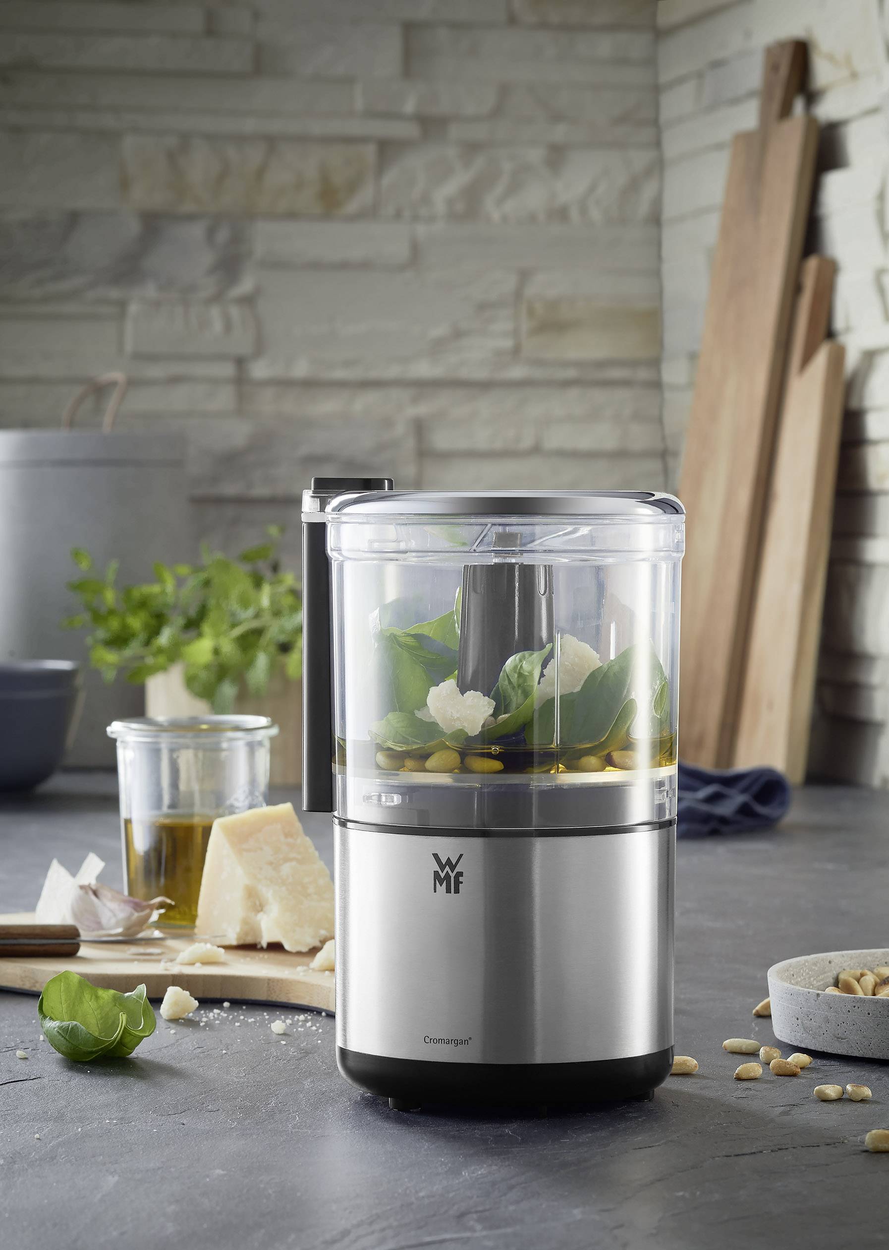 Silberner Küchenmixer auf einer grauen Arbeitsfläche, gefüllt mit Basilikum und Parmesan-Stückchen. Im Hintergrund Kräuter und Olivenöl.